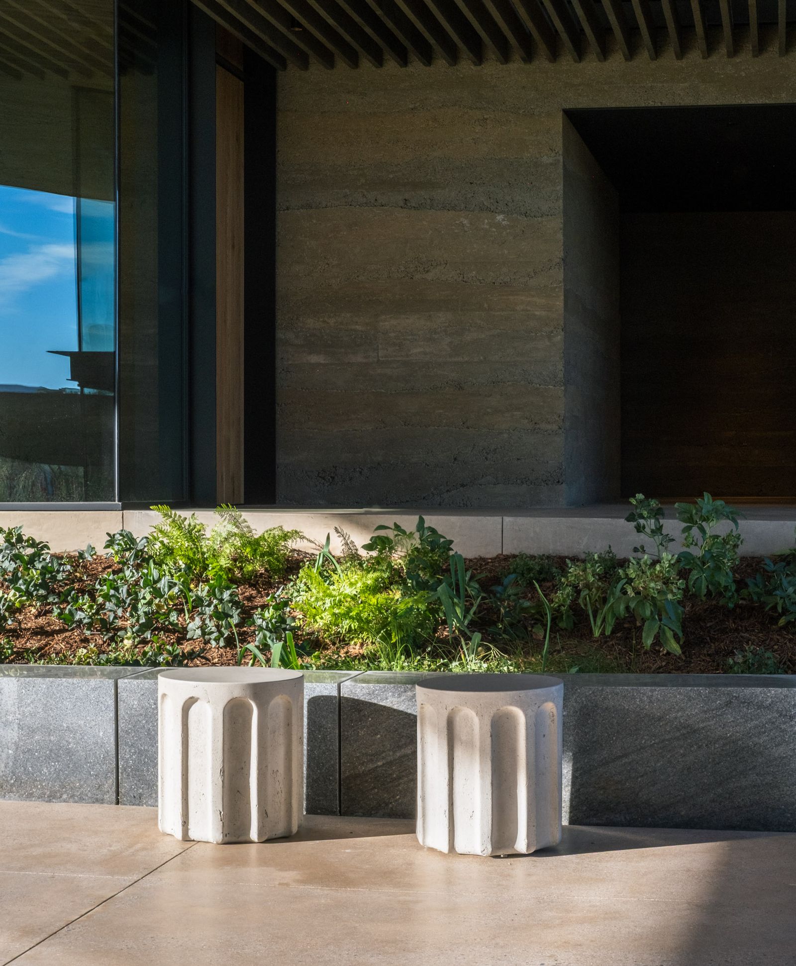 Contemporary outdoor space with two fluted concrete Forum side tables beside a landscaped planter and modern architectural facade.