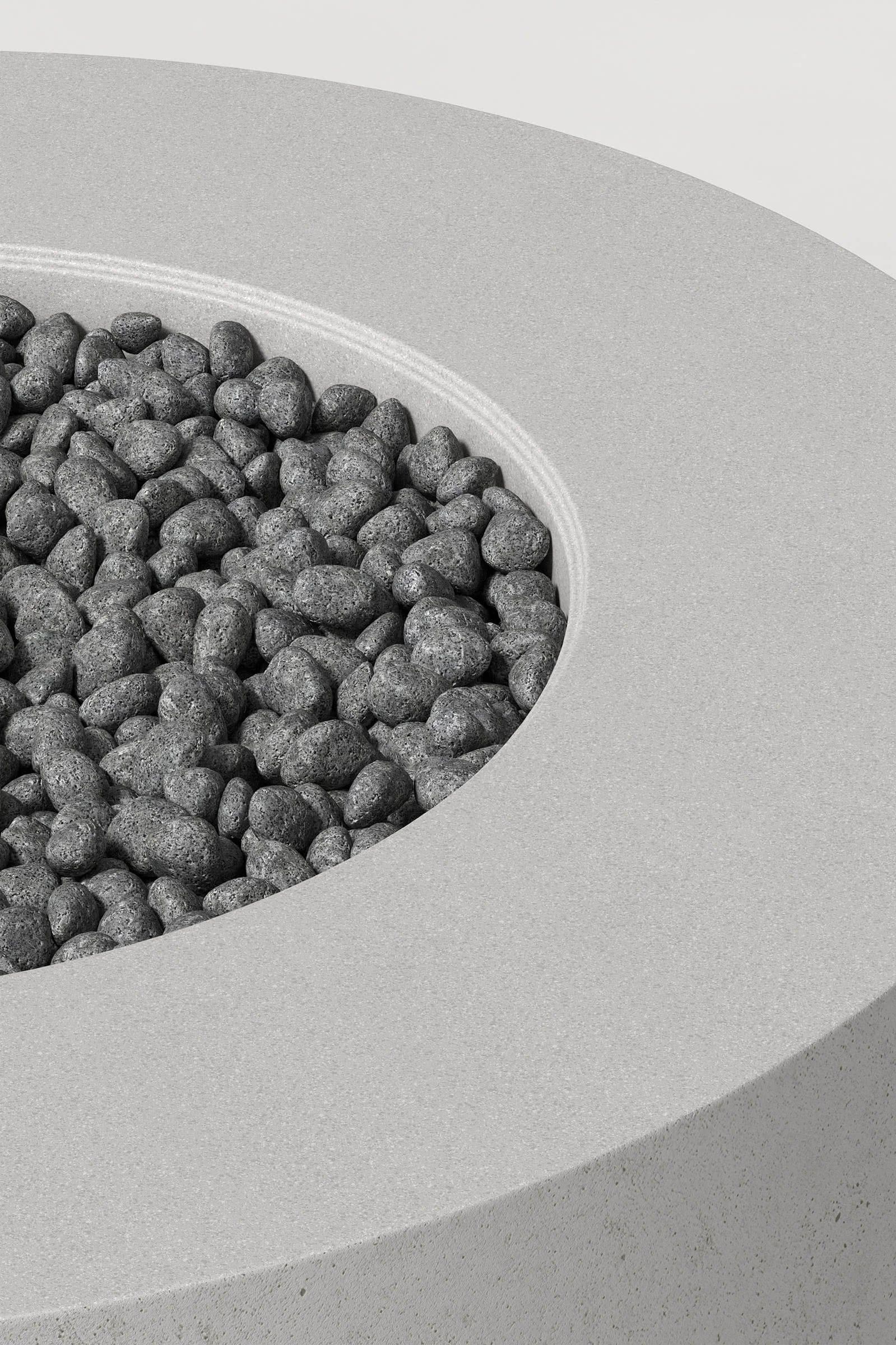 Close-up photograph of a cylindrical soft grey concrete fire pit with thick walls and a well filled with volcanic rocks. Showcases a smooth texture and precise edge design.
