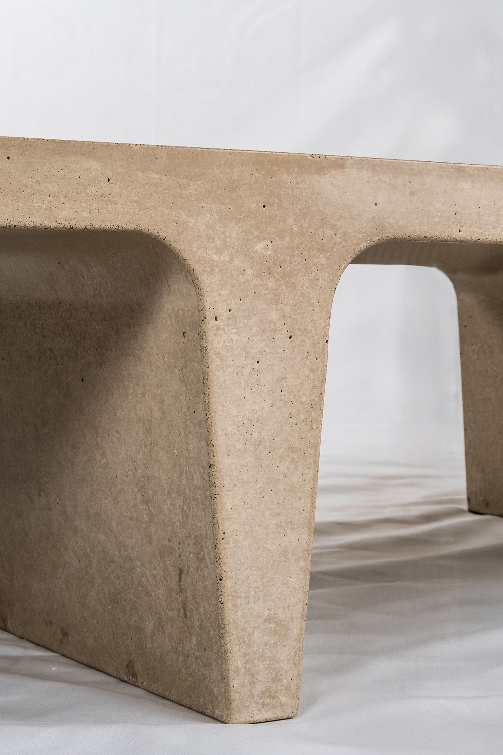Close-up of a light beige concrete coffee table with sculptural double tee legs, highlighting the troweled finish and softly rounded edges against a neutral backdrop.