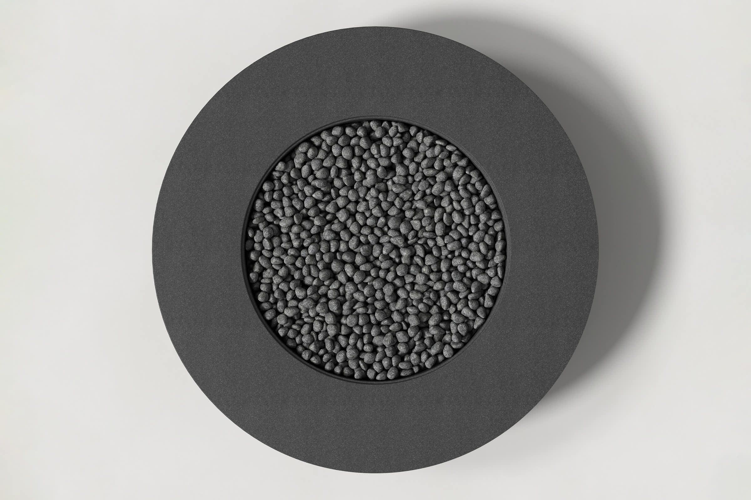 Top-down view of a circular charcoal concrete fire pit with thick walls and a circular well filled with volcanic rocks. Shown against a white background with slight shadows.