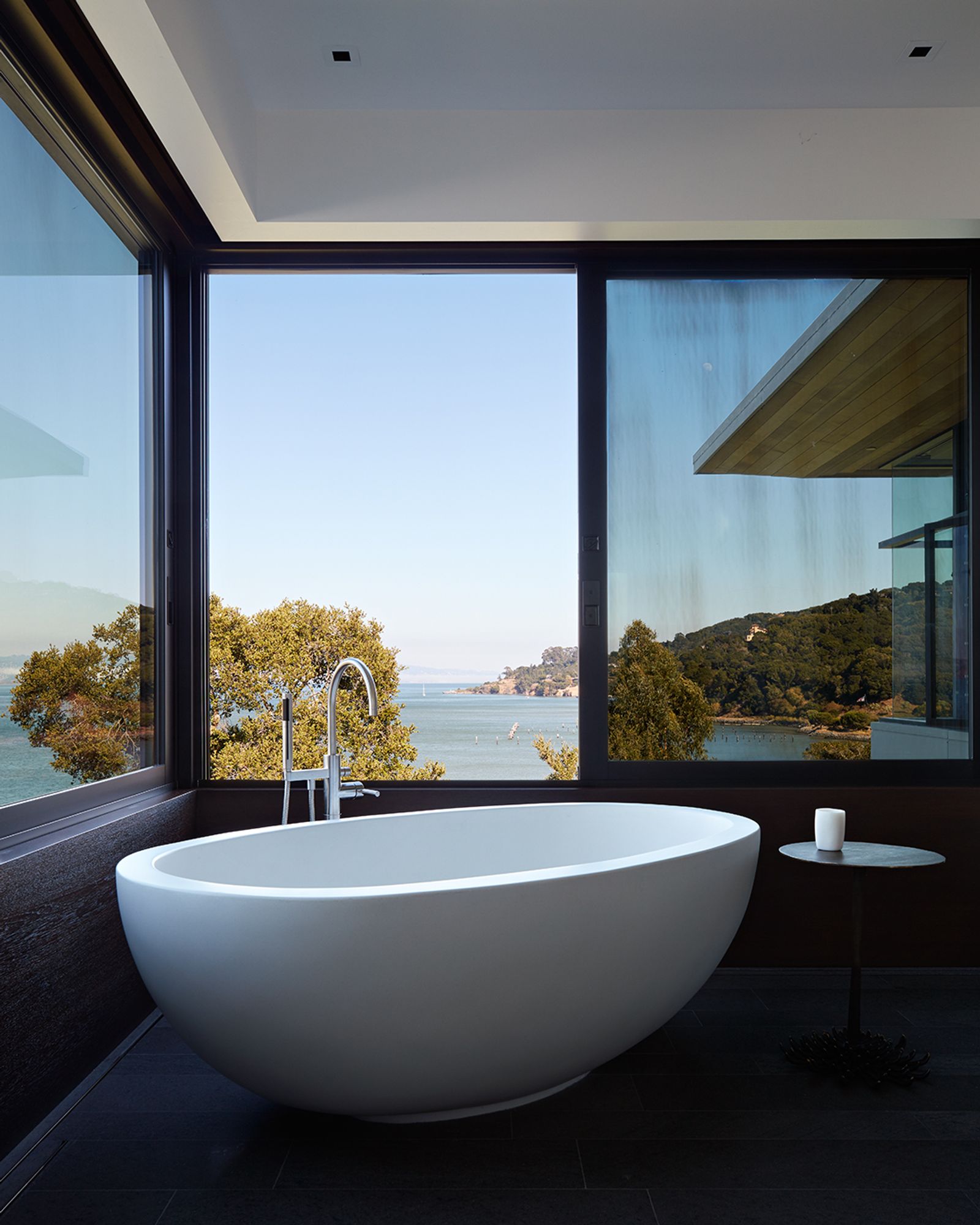 A modern freestanding white bathtub sits in a corner bathroom with floor-to-ceiling windows, offering expansive views of the ocean and surrounding trees. A small round side table with a candle is placed beside the tub.        Ask ChatGPT