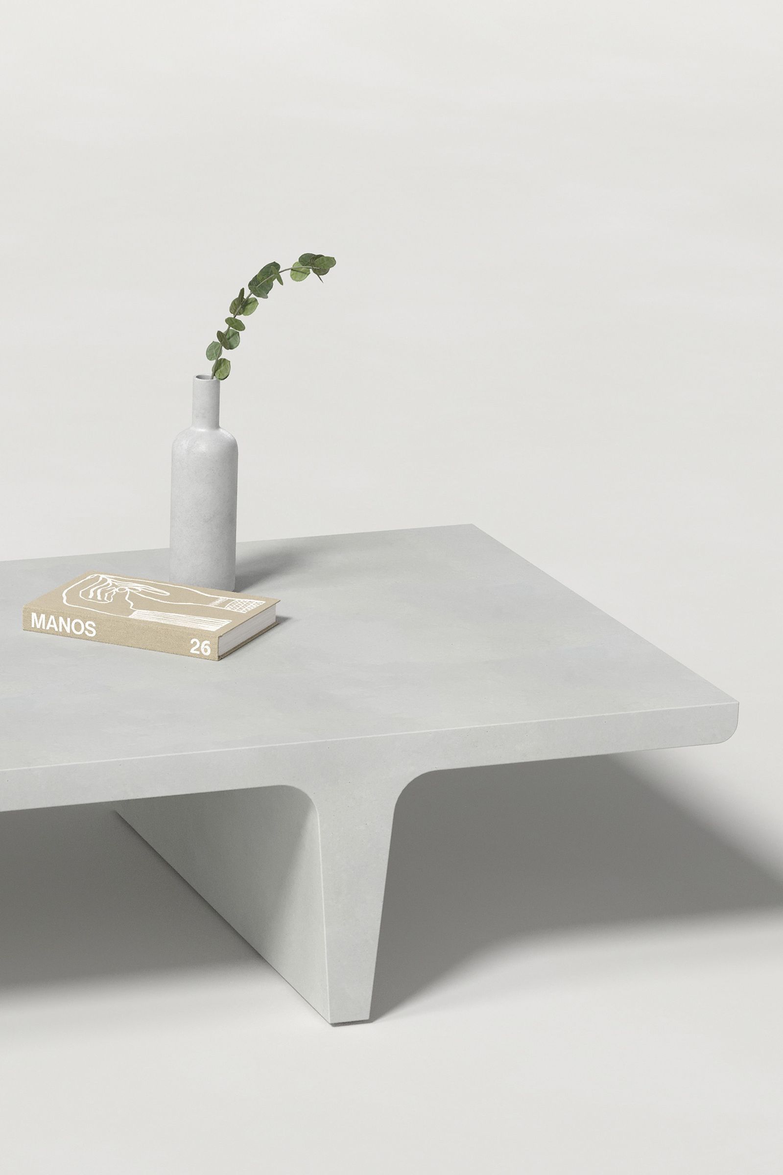 Close-up of a rectangular white concrete coffee table with sculptural double tee legs, styled with a neutral-toned book and a slender vase holding a eucalyptus, set against a clean white background.