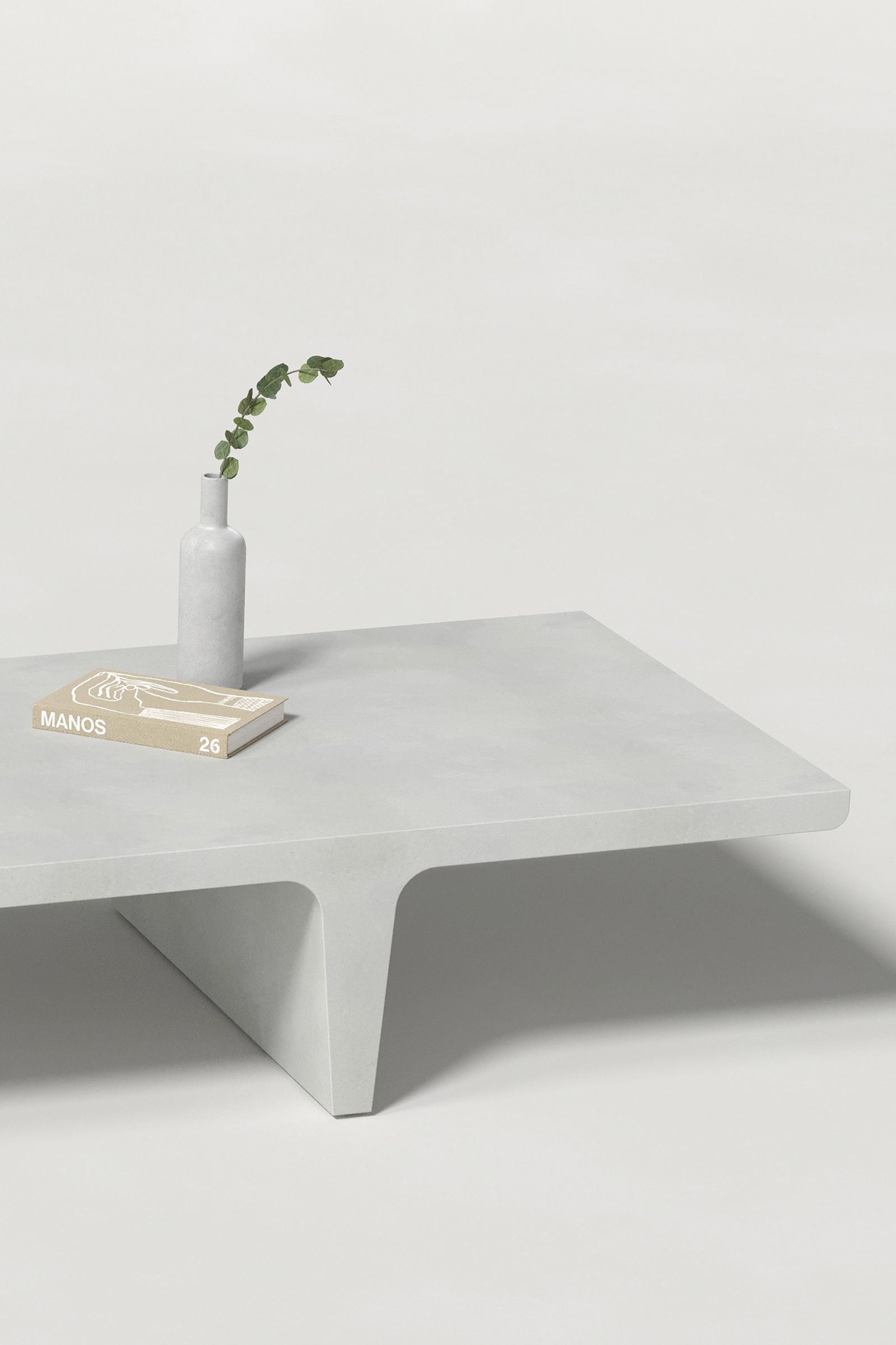 Close-up of a long rectangular white concrete coffee table with sculptural double tee legs, styled with a neutral-toned book and a slender vase holding a eucalyptus, set against a clean white background.