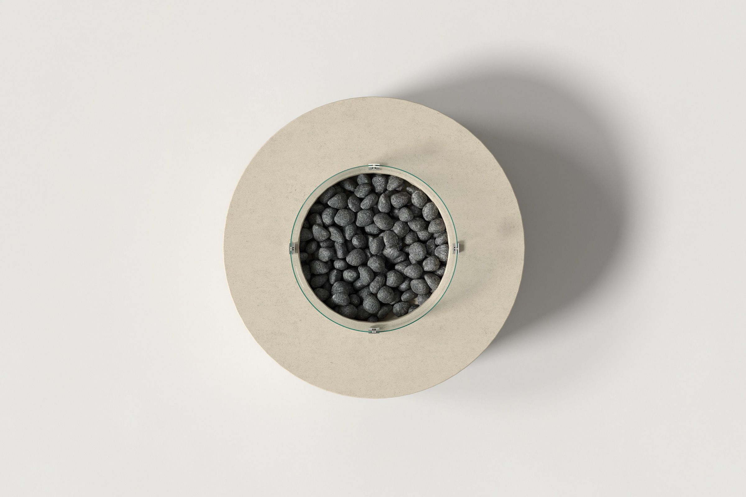 Top-down view of a circular sand-colored concrete fire pit with thick walls and a central well filled with dark volcanic rocks, encircled by a clear glass shield on a white background.