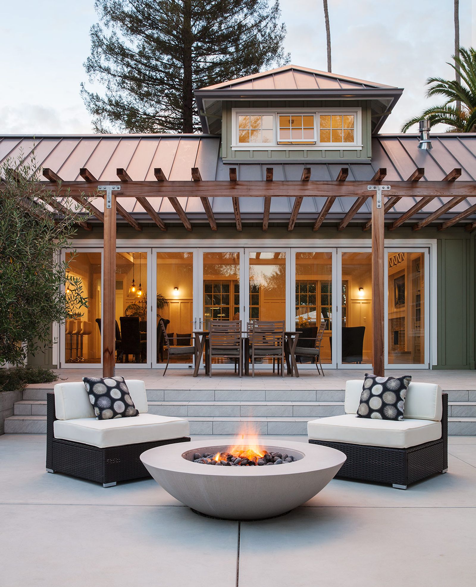 A backyard patio with a lit concrete fire pit between two outdoor chairs. Backgrounded by a wooden pergola and floor-to-ceiling glass doors up three steps, leading into a warmly lit interior.
