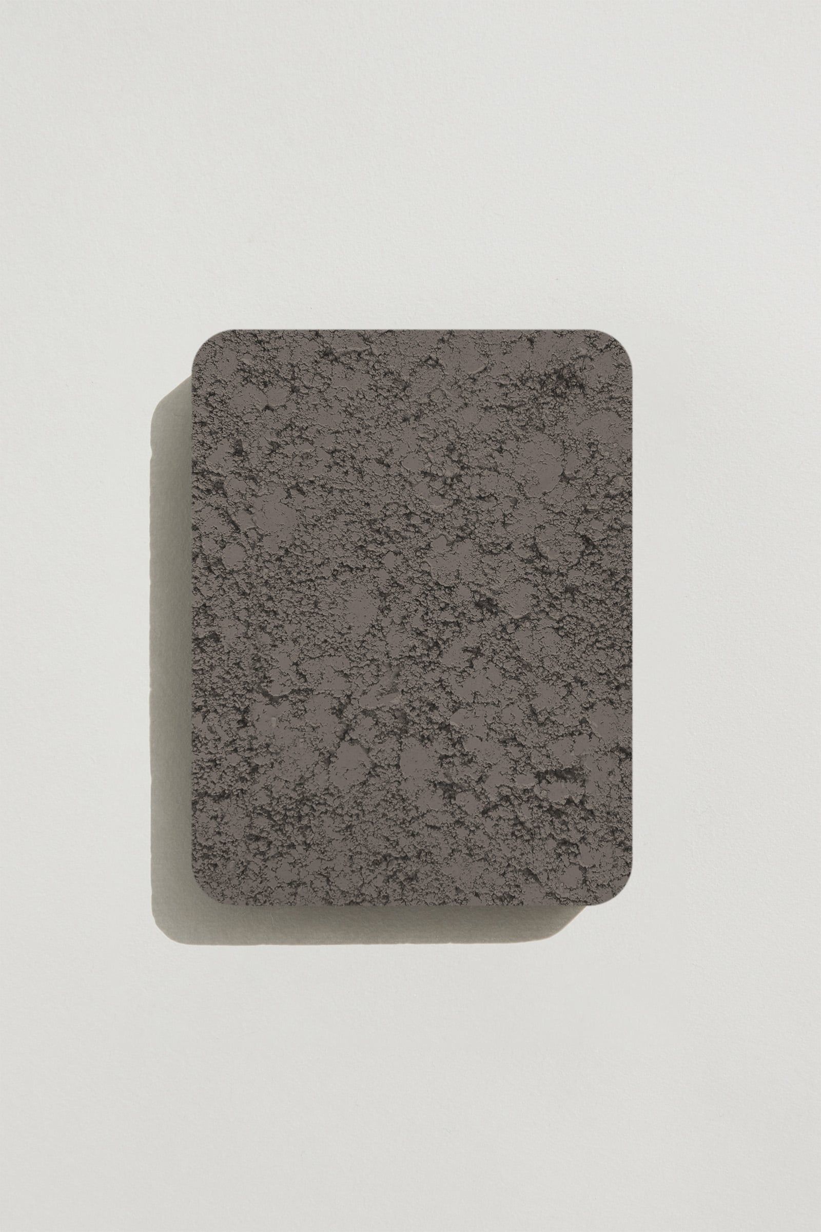 A rectangular sample of rough dark grey concrete with a natural, pitted surface texture. Photographed on a white background with a cast shadow.