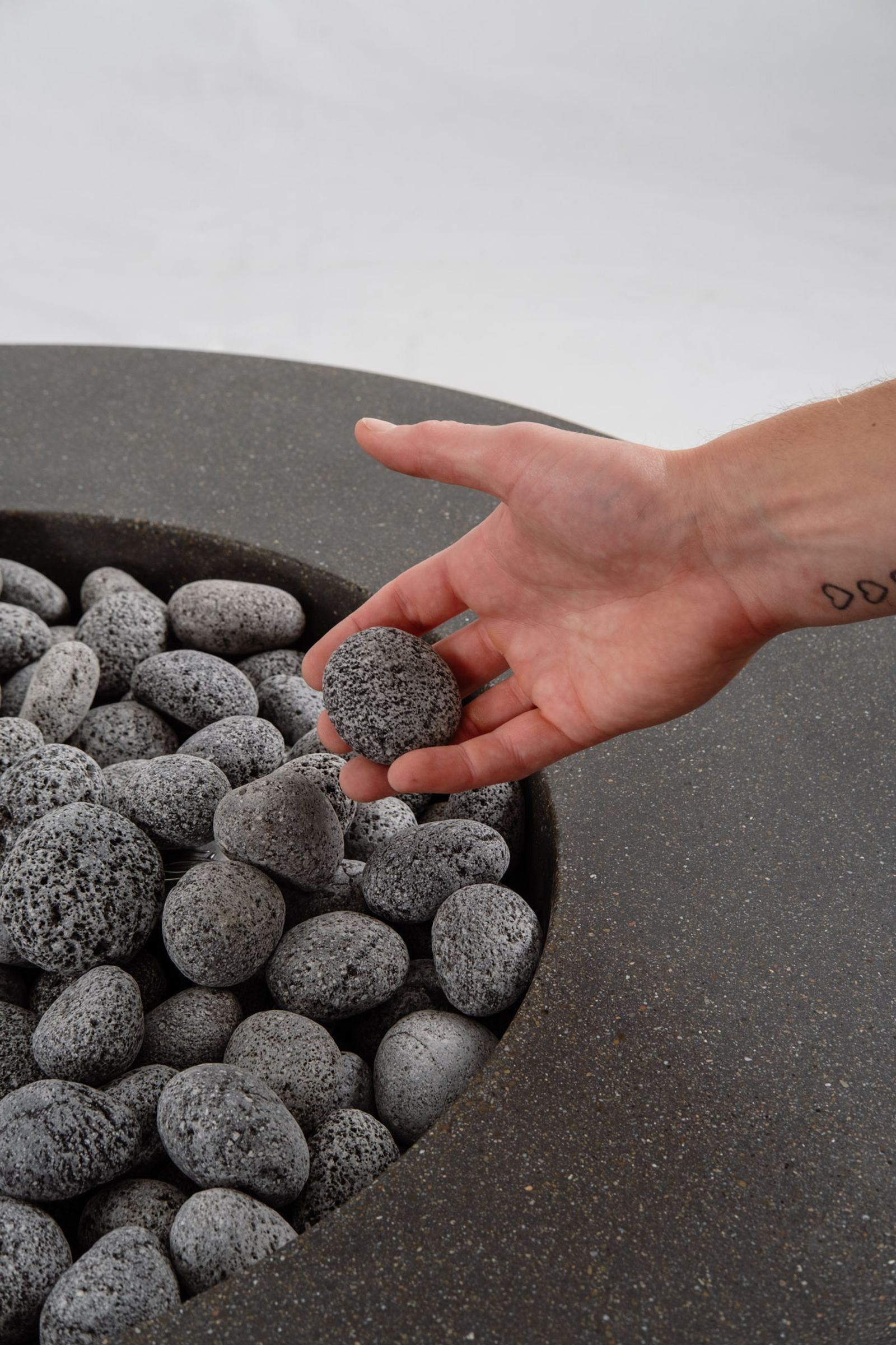 Close-up of the Rosa a round dark grey concrete fire pit with a honed top and lava rocks, with a hand holding the lava rock over it.