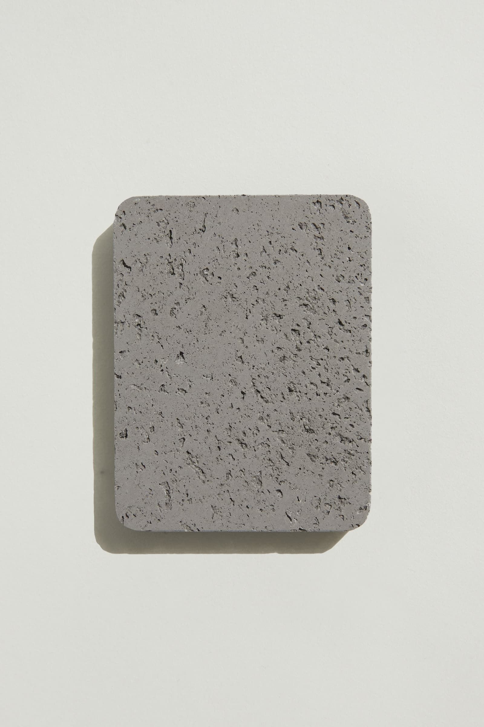 A rectangular sample of rough grey concrete with a natural, pitted surface texture. Photographed on a white background with a cast shadow.