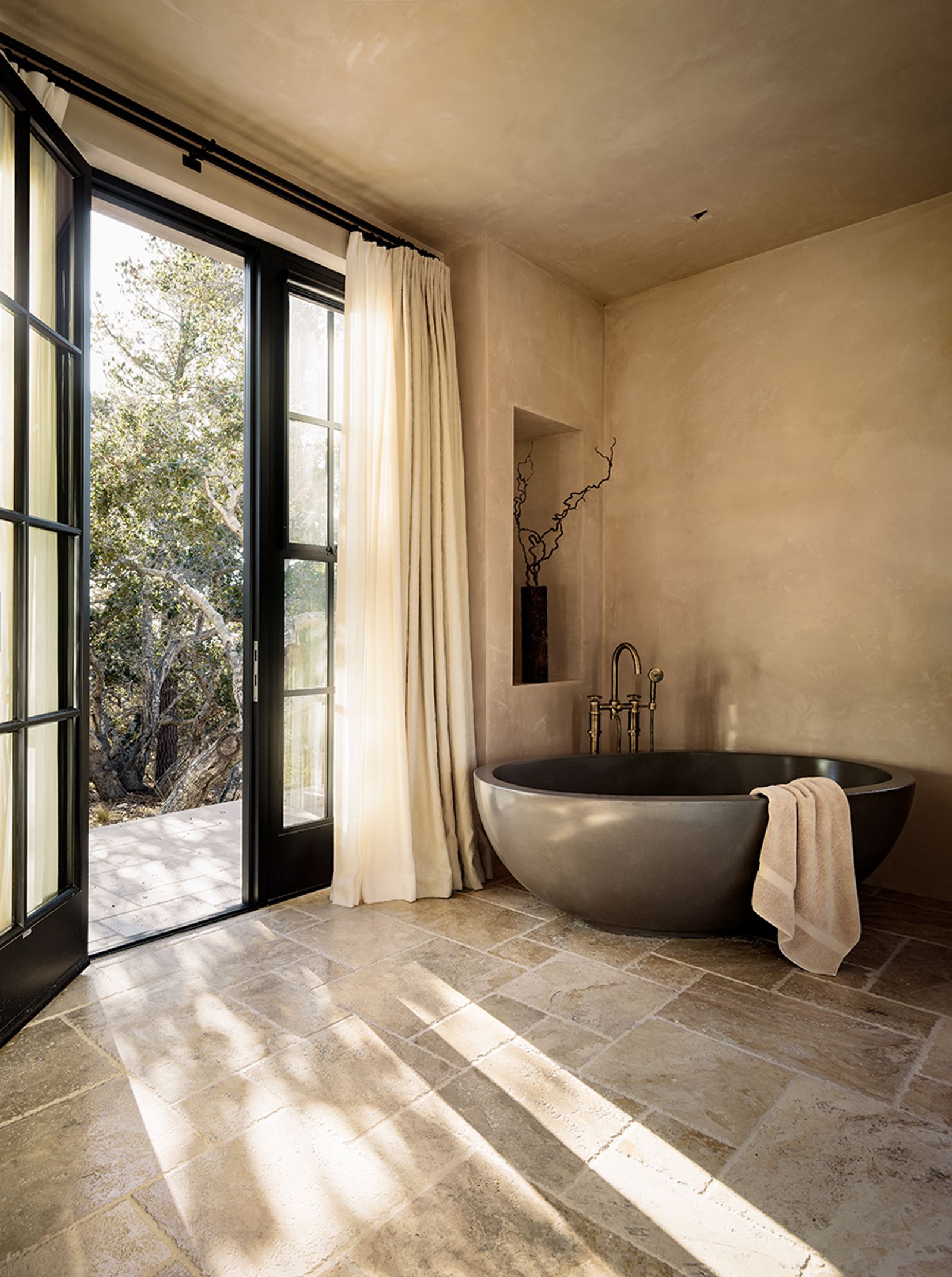A dark, round freestanding bathtub sits in a sunlit bathroom with natural stone floors and plastered walls. French doors open to a wooded outdoor area, and soft curtains frame the view. A towel drapes over the edge of the tub, and a decorative vase with branches rests in a wall niche above the faucet.