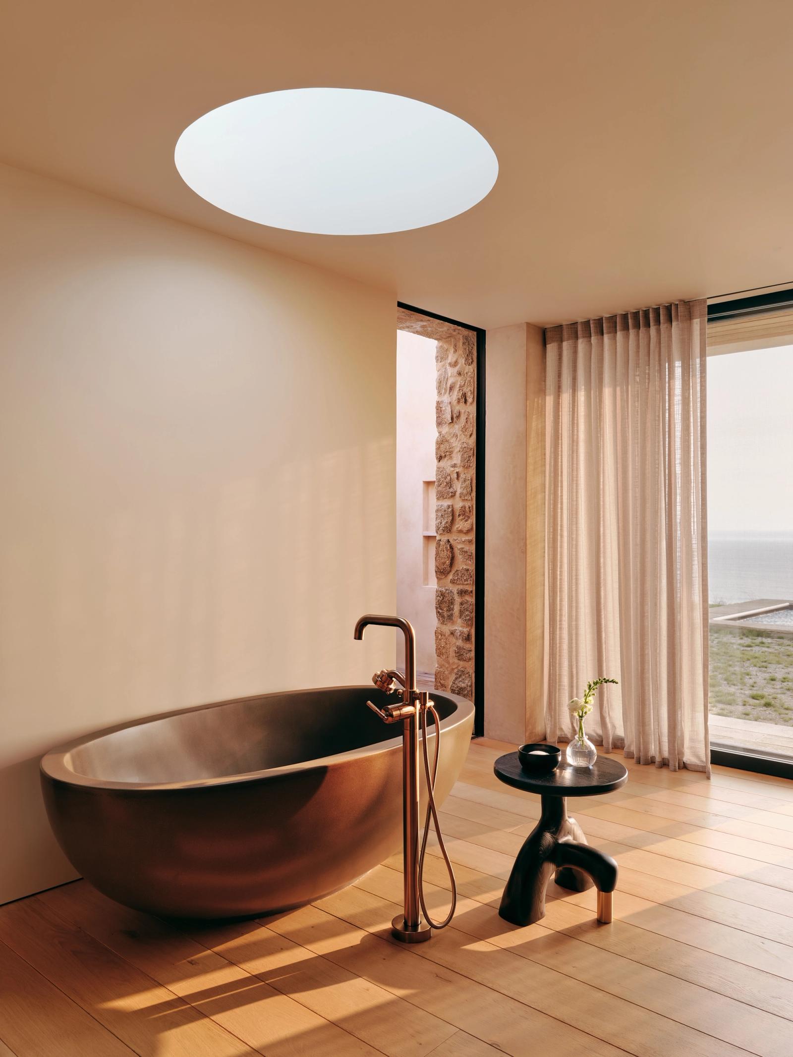 Luxurious bathroom with bronze freestanding oval tub, round skylight, and sheer curtains. Warm wood floors and stone wall detail create spa-like atmosphere.