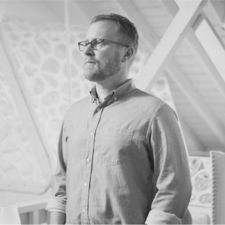 Black and white photo of a man with short hair, beard and glasses wearing a button-up shirt, looking to the side with geometric patterns in the background.