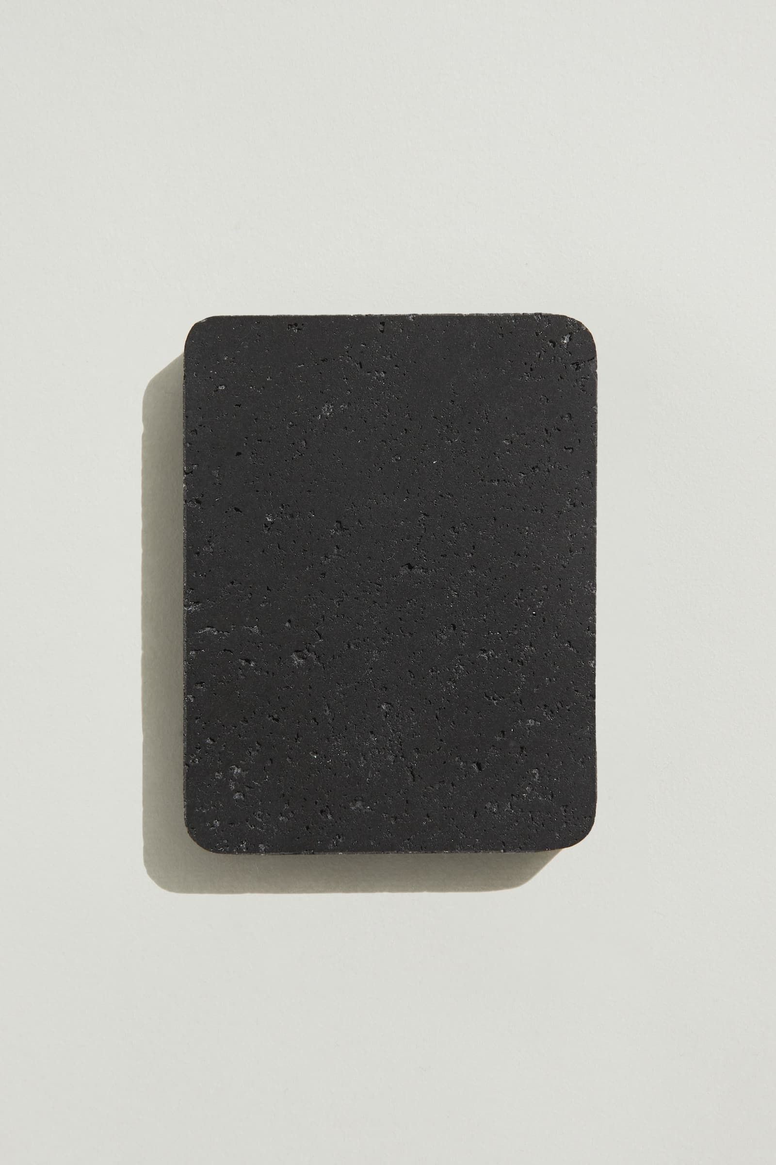 A rectangular sample of rough jet black concrete with a natural, pitted surface texture. Photographed on a white background with a cast shadow.