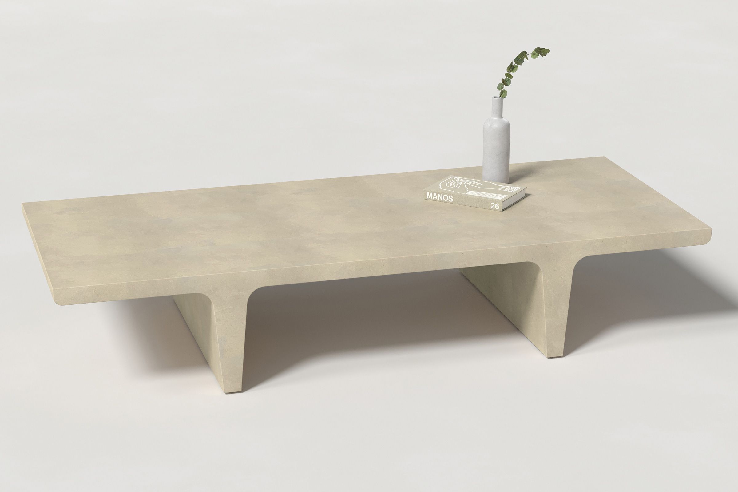 A rectangular light beige concrete coffee table with sculptural double tee legs, styled with a neutral-toned book and a slender vase holding a eucalyptus, set against a clean white background.