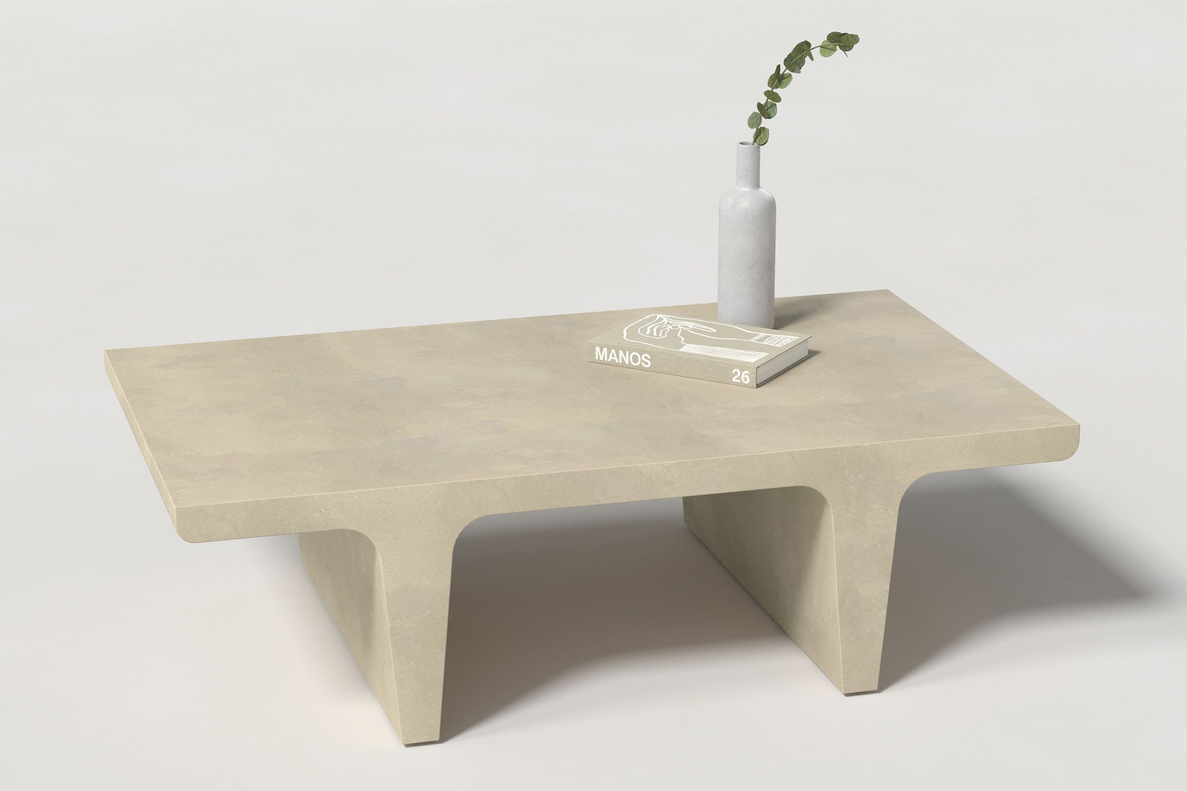 A rectangular light beige concrete coffee table with sculptural double tee legs, styled with a neutral-toned book and a slender vase holding a eucalyptus, set against a clean white background.