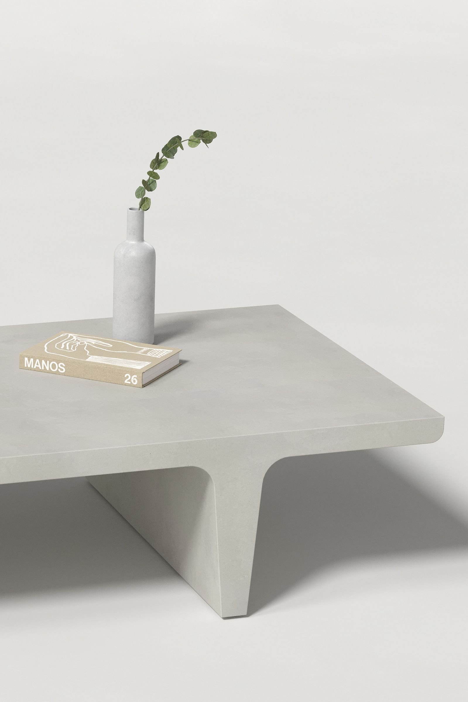 Close-up of a rectangular light grey concrete coffee table with sculptural double tee legs, styled with a neutral-toned book and a slender vase holding a eucalyptus, set against a clean white background.