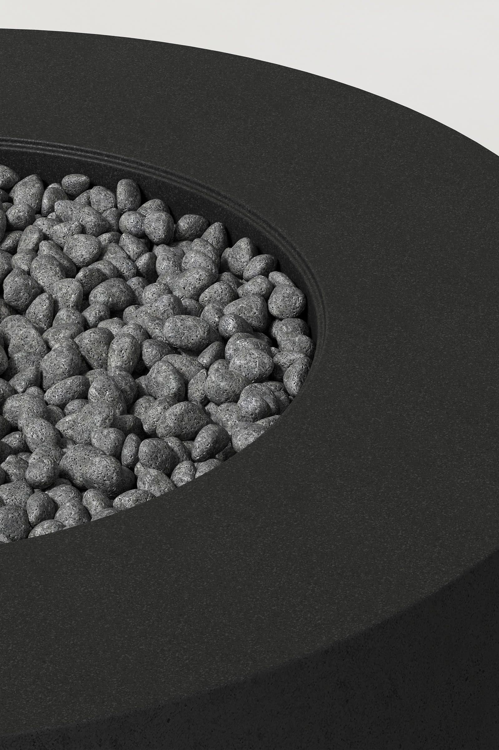 Close-up photograph of a cylindrical jet black concrete fire pit with thick walls and a well filled with volcanic rocks. Showcases a smooth texture and precise edge design.