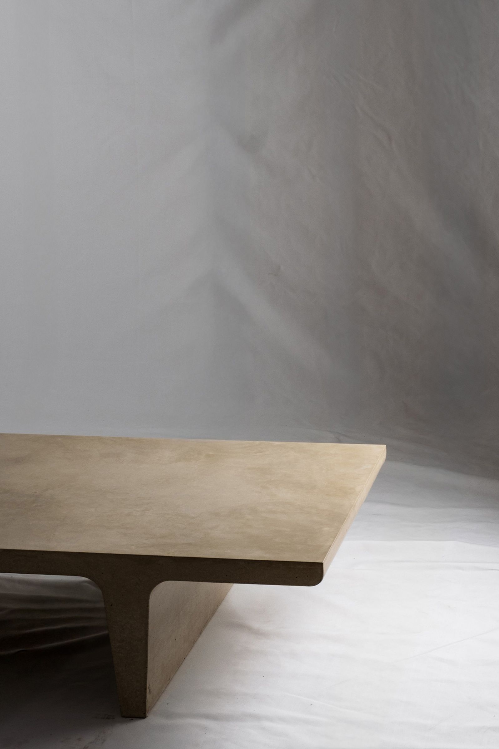 A close up of a light beige concrete coffee table with sculptural double tee legs, featuring a smooth troweled finish, set against a soft neutral backdrop.