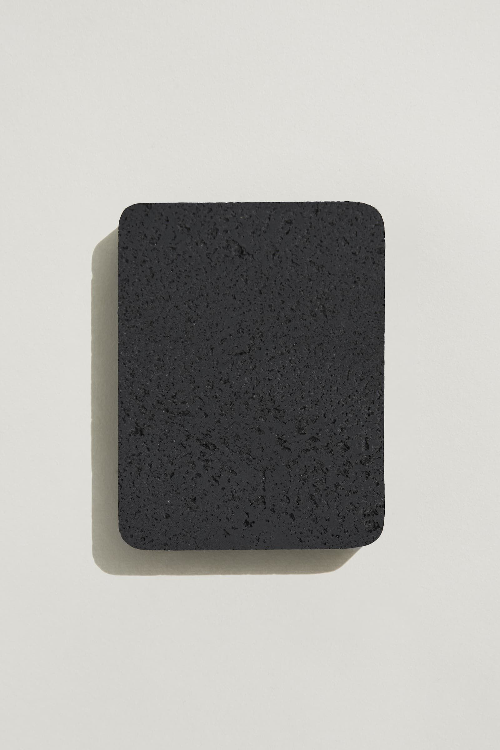 A rectangular sample of rough charcoal concrete with a natural, pitted surface texture. Photographed on a white background with a cast shadow.