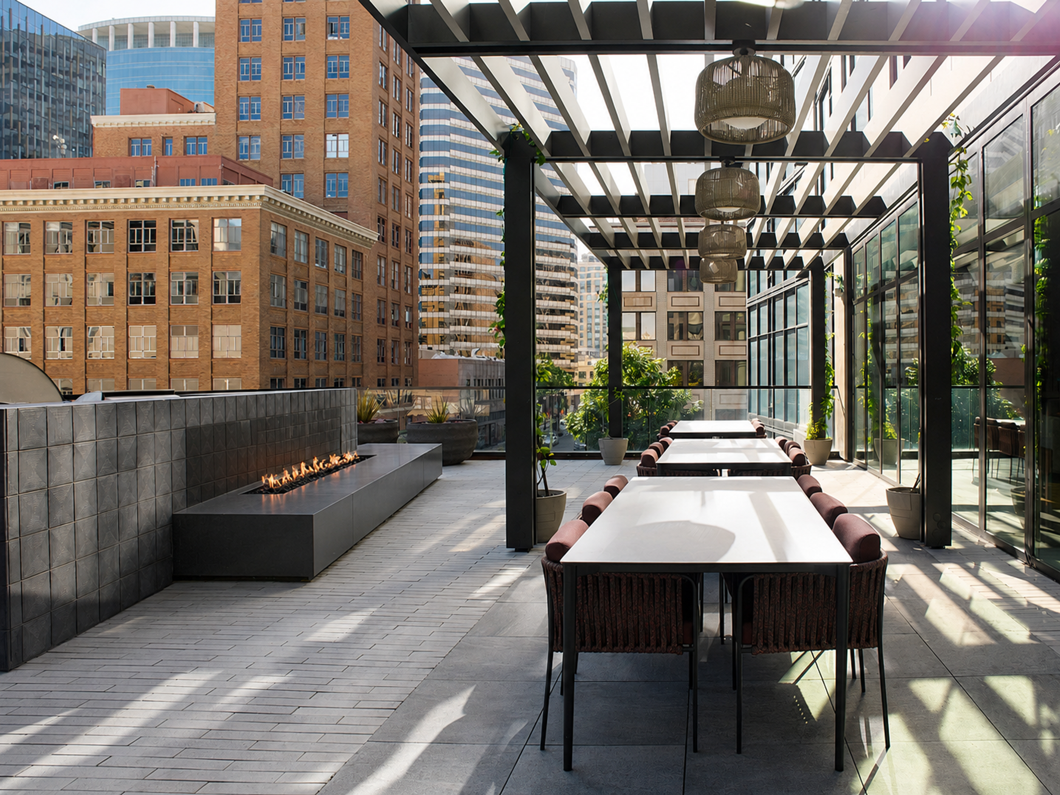 A rooftop dining area with long tables and brown chairs. Hanging woven lamps hang down, with city views of brick buildings in the background.