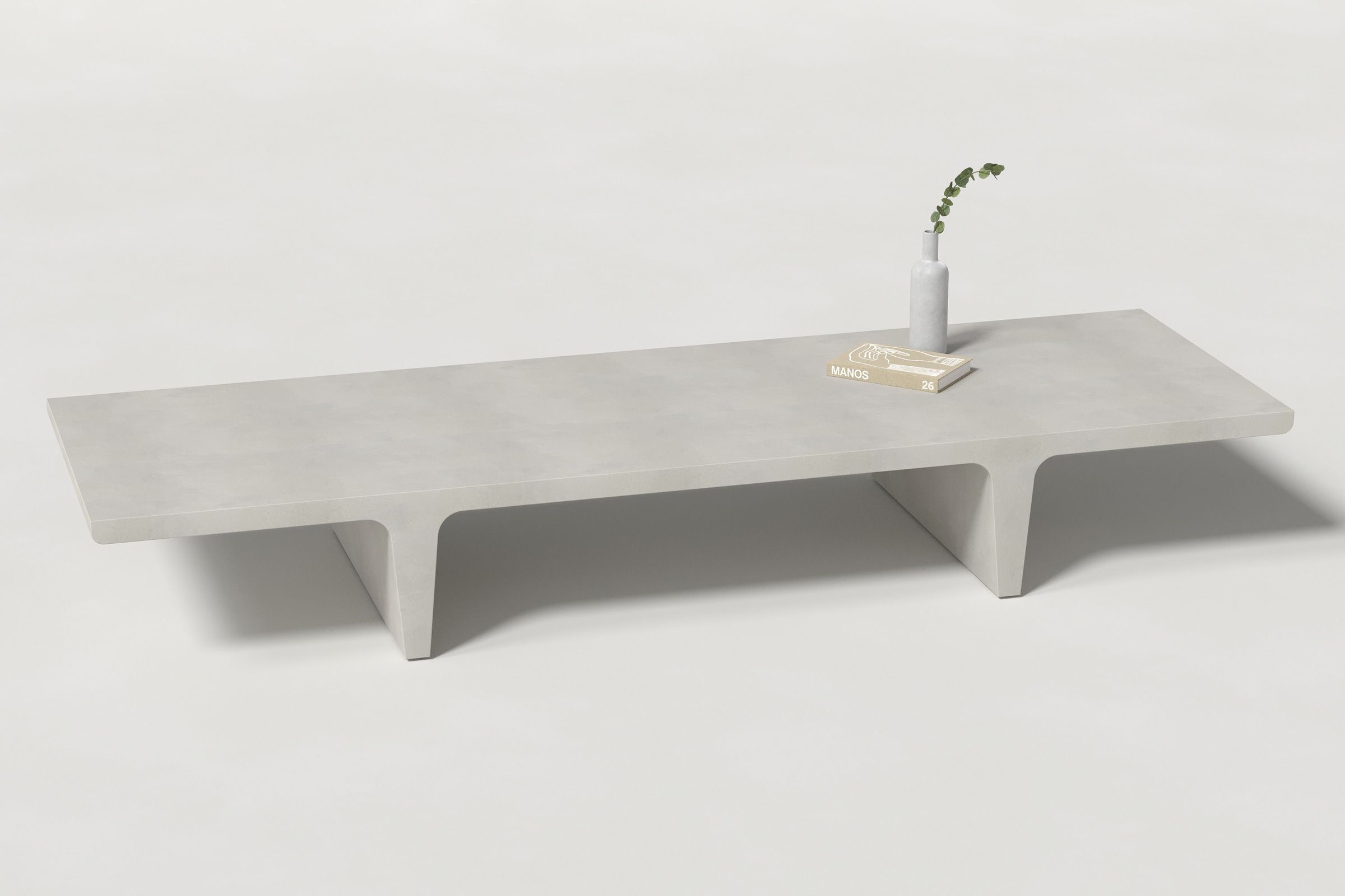 A long rectangular light grey concrete coffee table with sculptural double tee legs, styled with a neutral-toned book and a slender vase holding a eucalyptus, set against a clean white background.