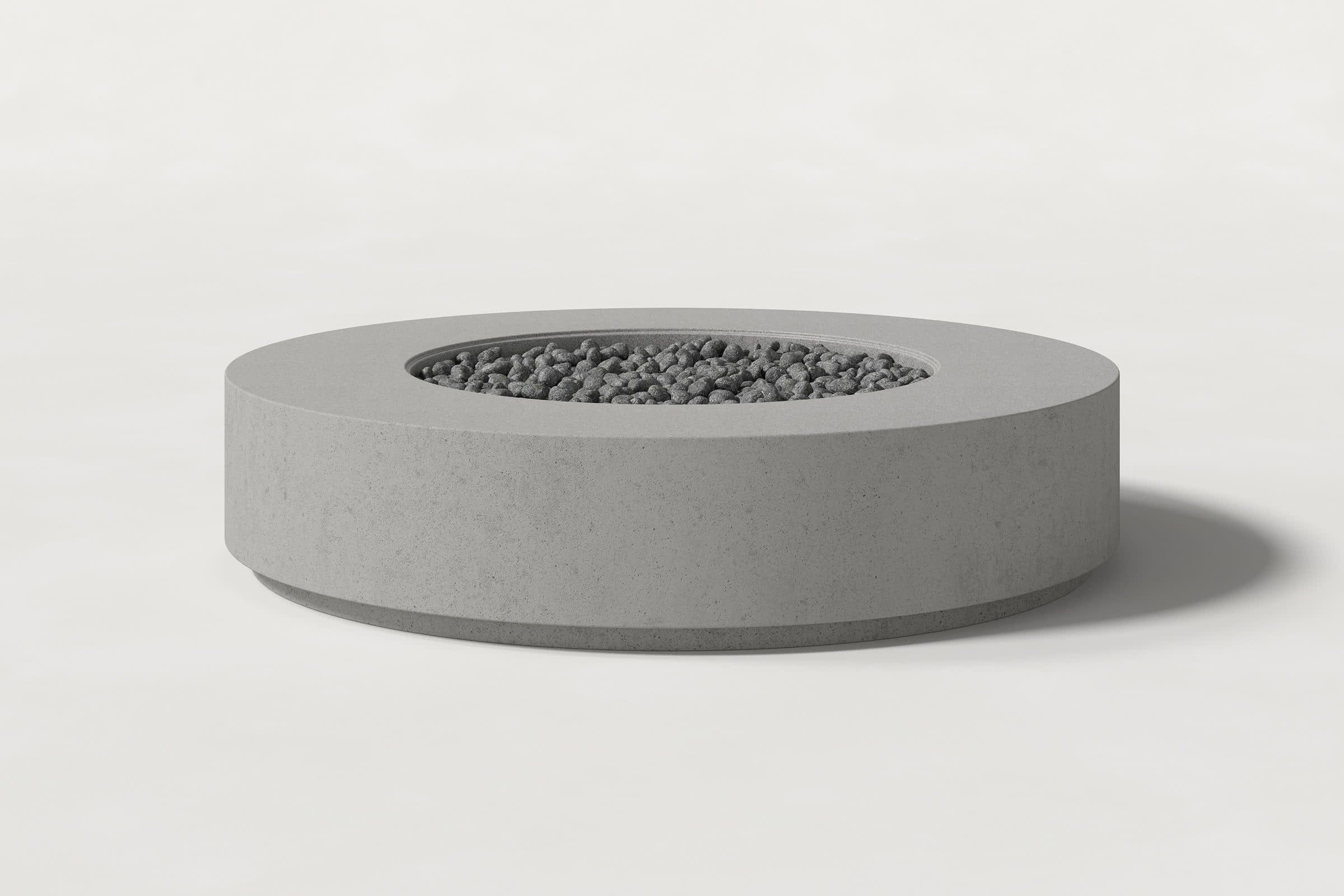 Low cylindrical grey concrete fire pit with thick walls and a central circular well filled with dark volcanic rocks. Side view on a white background with a shadow to the right.