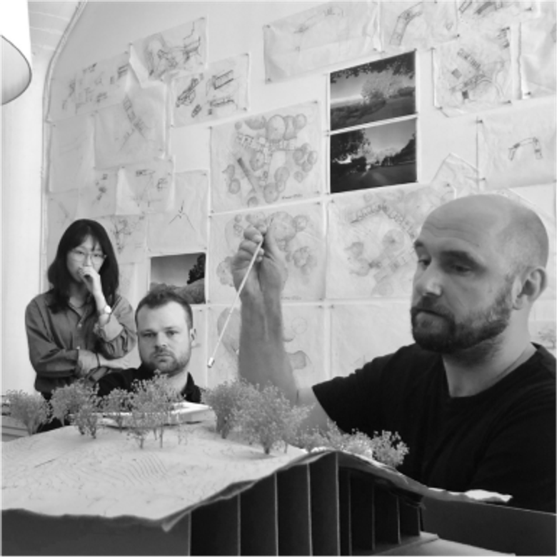 Black and white photo of three people in a workspace with architectural models and drawings on walls. A man points at a model while others observe.