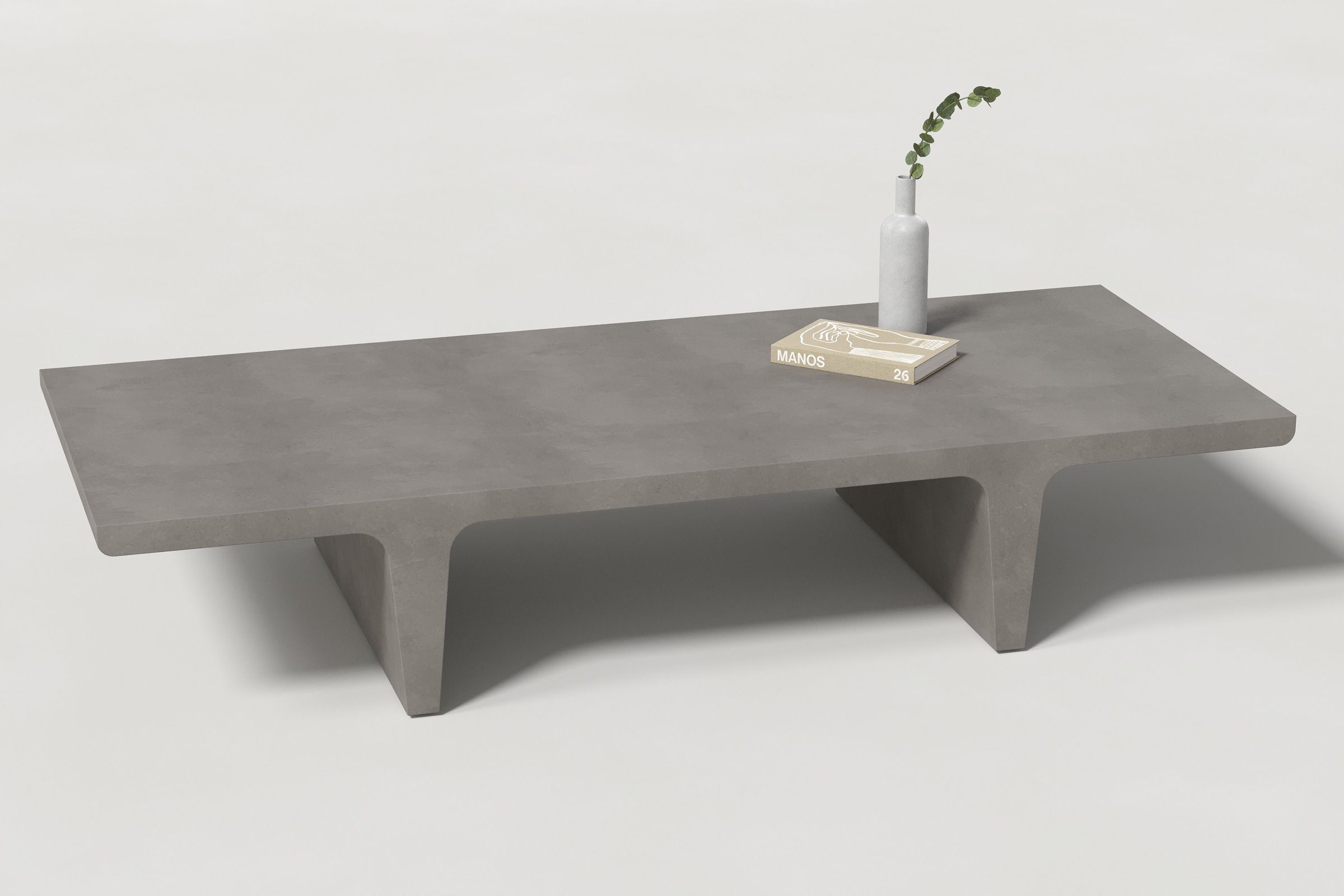 A rectangular dark grey concrete coffee table with sculptural double tee legs, styled with a neutral-toned book and a slender vase holding a eucalyptus, set against a clean white background.