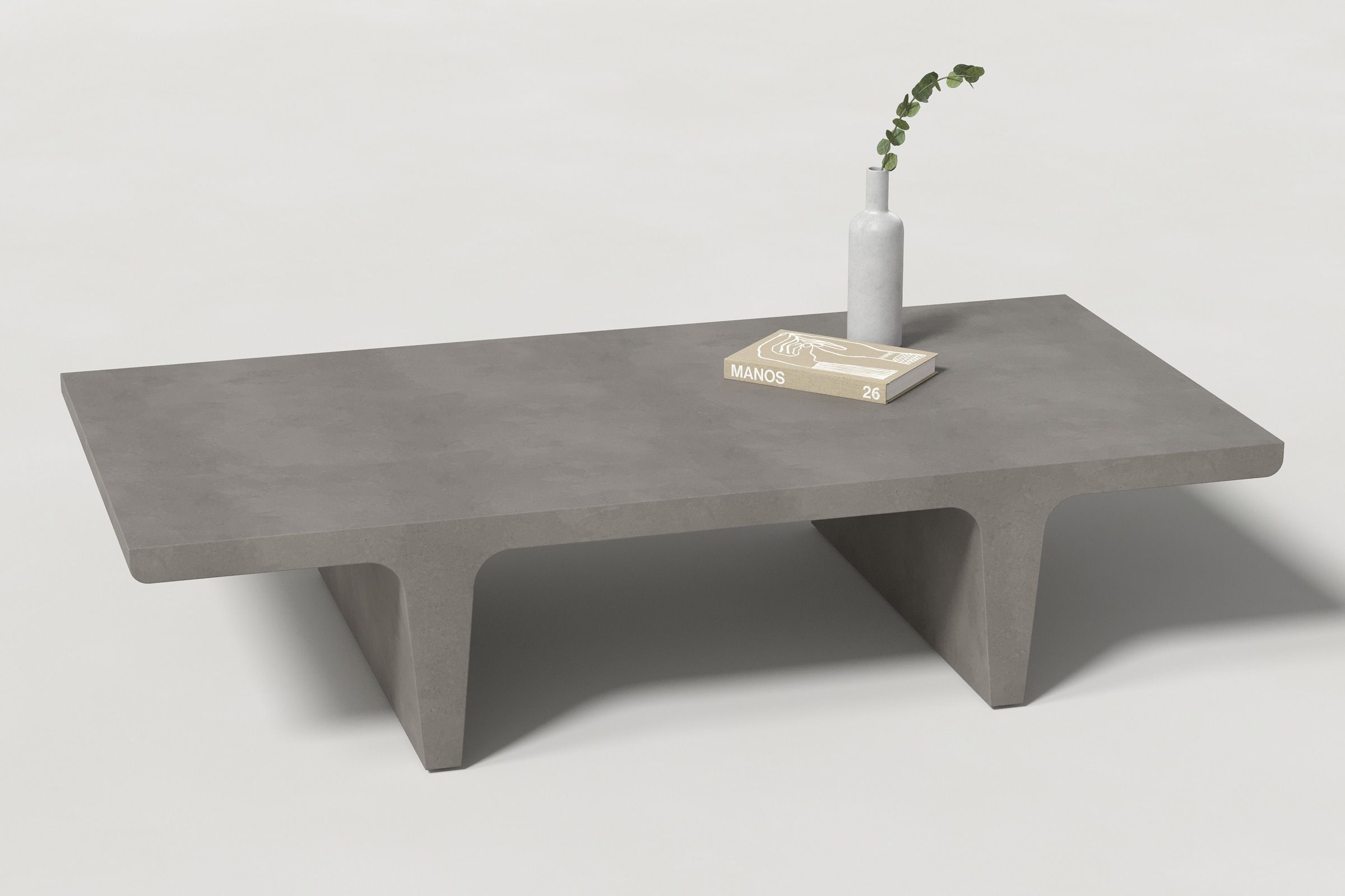 A rectangular dark grey concrete coffee table with sculptural double tee legs, styled with a neutral-toned book and a slender vase holding a eucalyptus, set against a clean white background.