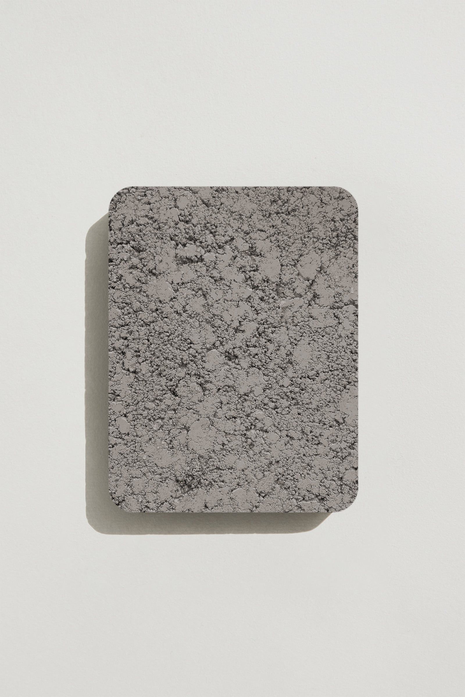 A rectangular sample of rough grey concrete with a natural, pitted surface texture. Photographed on a white background with a cast shadow.
