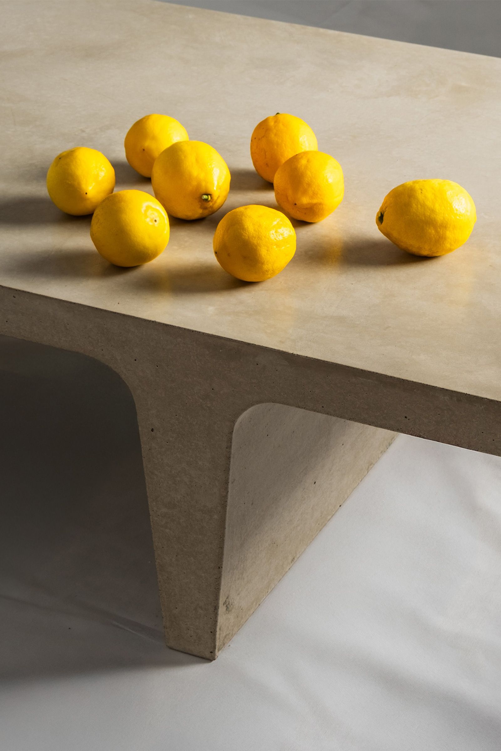 Close-up of a light beige concrete coffee table with sculptural double tee legs, featuring a smooth top with scattered yellow lemons against a neutral backdrop.