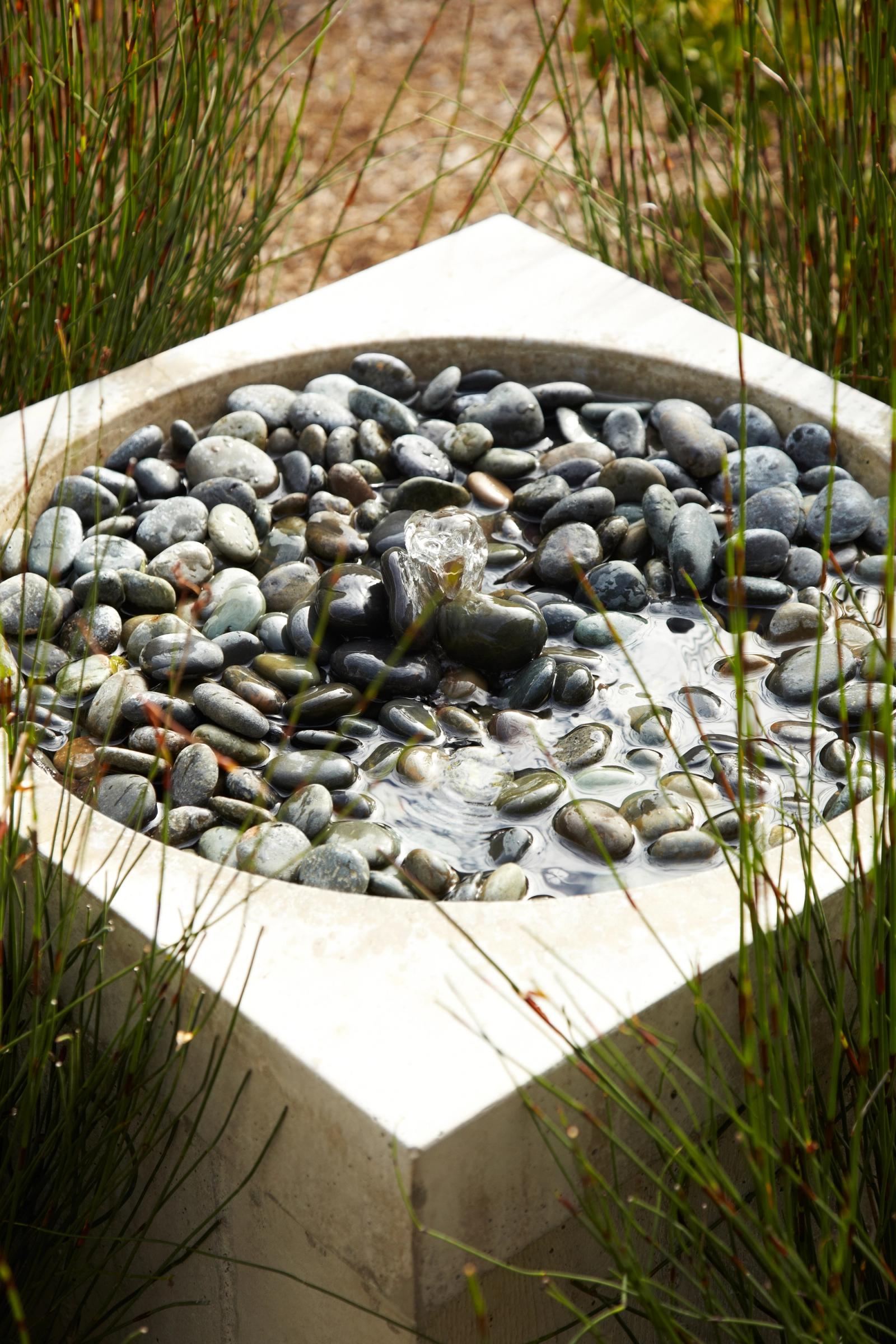 Square concrete water basin filled with smooth river stones, surrounded by tall grasses in a landscaped garden setting.