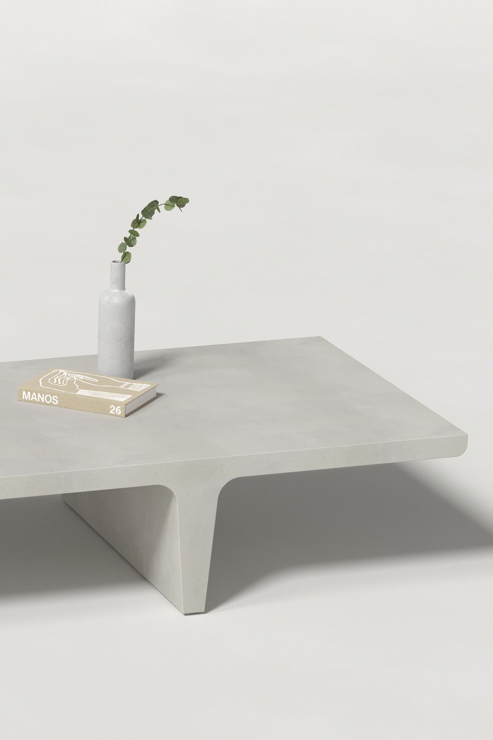 Close-up of a long rectangular light grey concrete coffee table with sculptural double tee legs, styled with a neutral-toned book and a slender vase holding a eucalyptus, set against a clean white background.