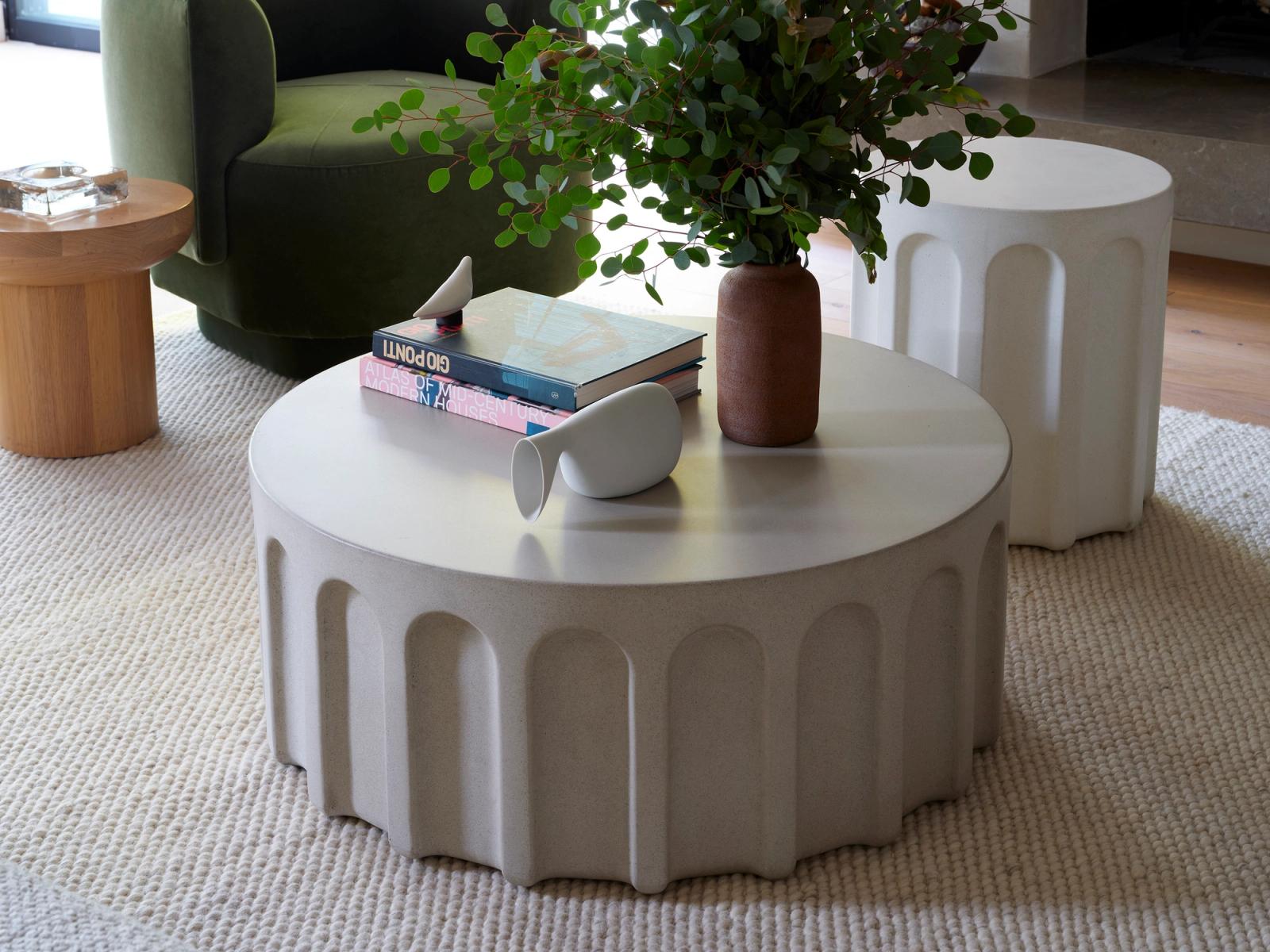 Large fluted white concrete coffee table in living room, styled with books and brown ceramic vase with foliage. Green velvet chair and textured carpet visible.