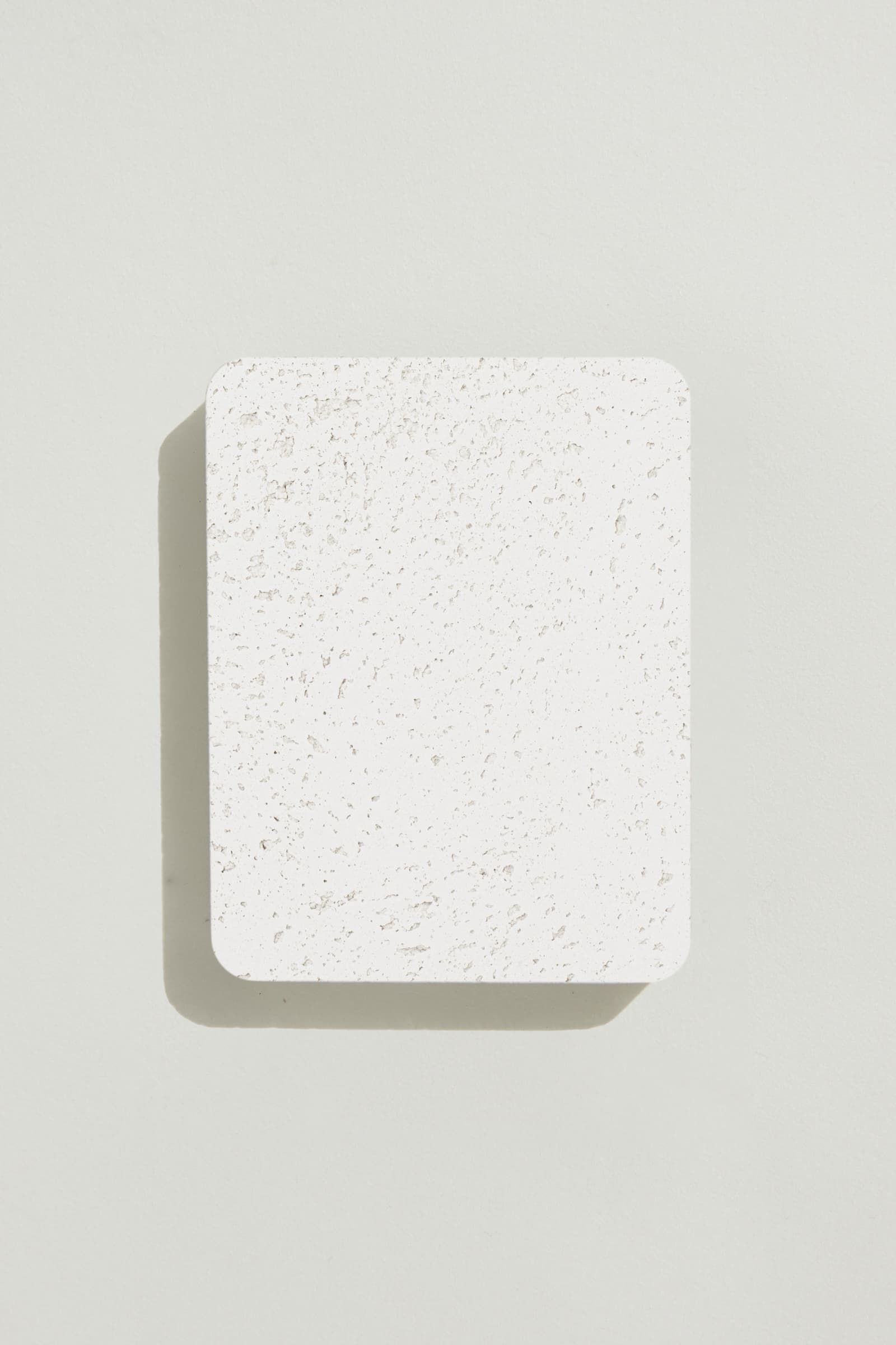 A rectangular sample of rough white concrete with a natural, pitted surface texture. Photographed on a white background with a cast shadow.