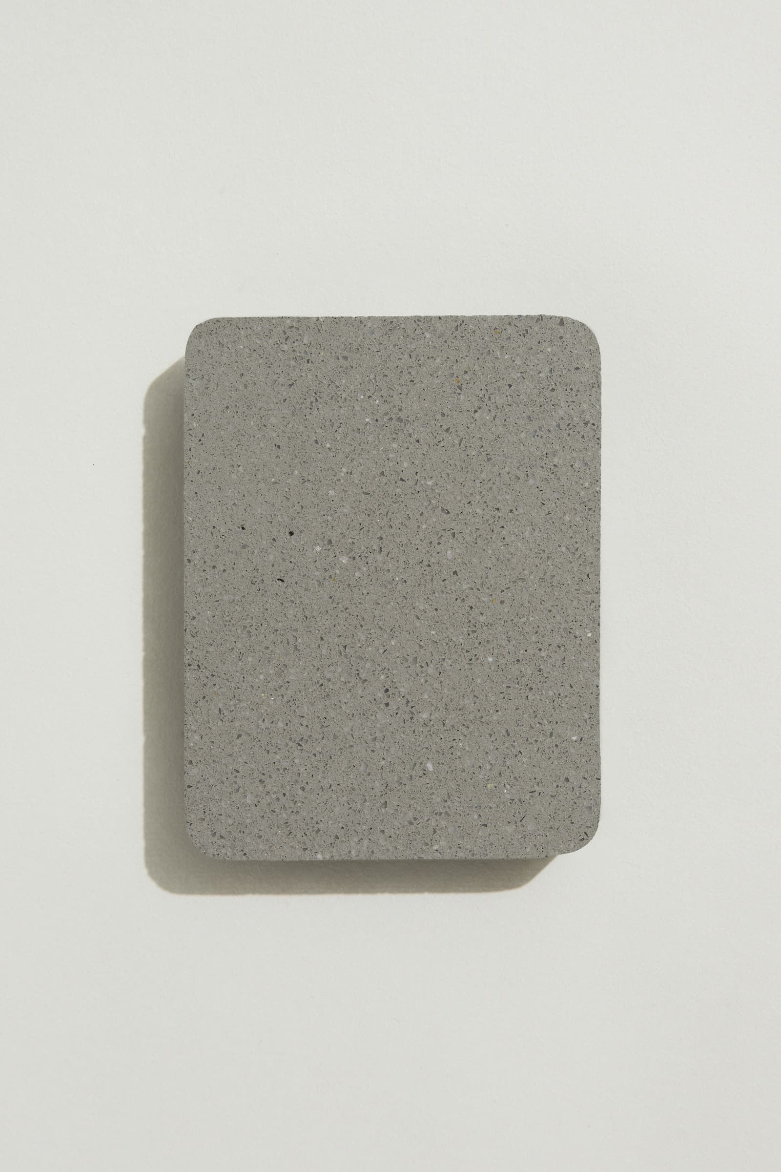 A rectangular sample of smooth grey concrete with grey, white and black terrazzo speckling, and rounded corners. Photographed on a white background with a cast shadow