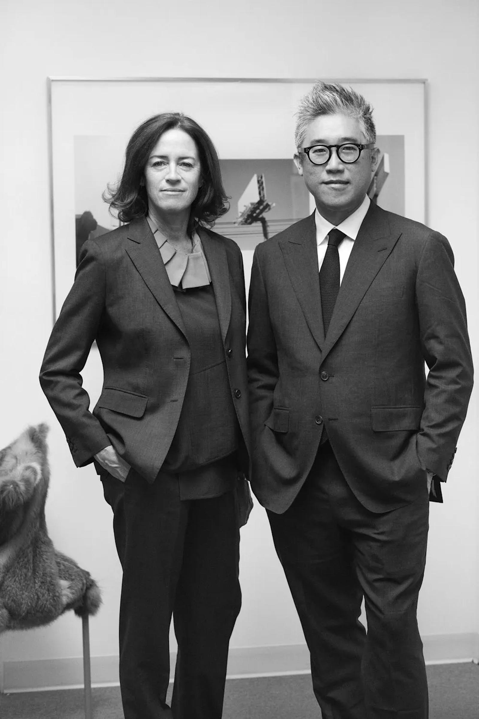 A black and white portrait of two people standing against a light wall. Both wear formal suits and project a professional demeanor. Artwork is partially visible behind them.