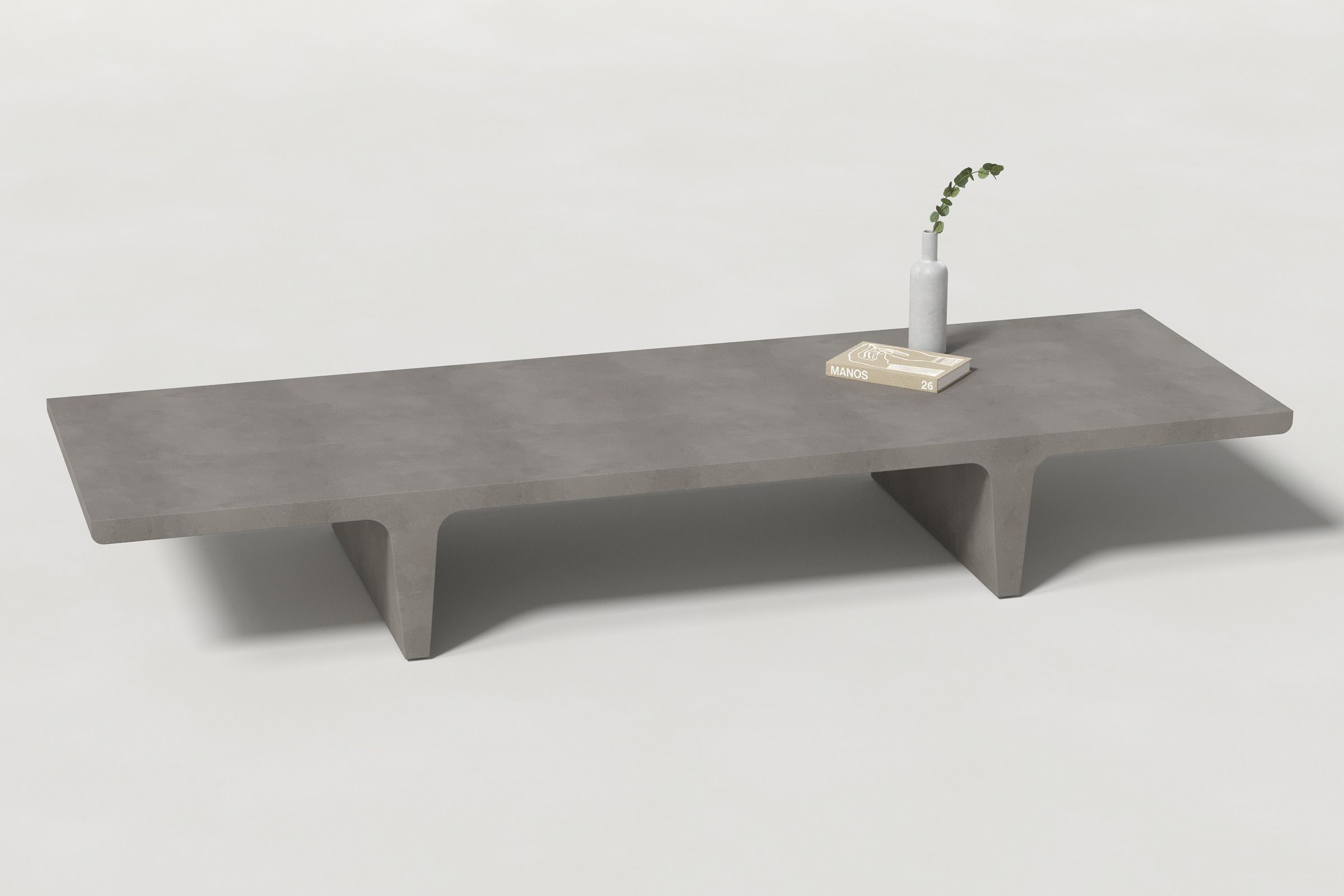 A long rectangular dark grey concrete coffee table with sculptural double tee legs, styled with a neutral-toned book and a slender vase holding a eucalyptus, set against a clean white background.