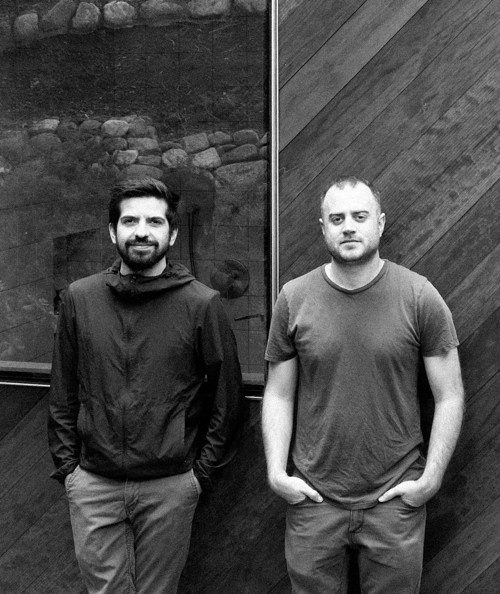 A black and white photograph of two people standing against dark contrasting stone & wooden wooden background. Both are wearing casual attire, one in a long-sleeved shirt , the other in a t-shirt.