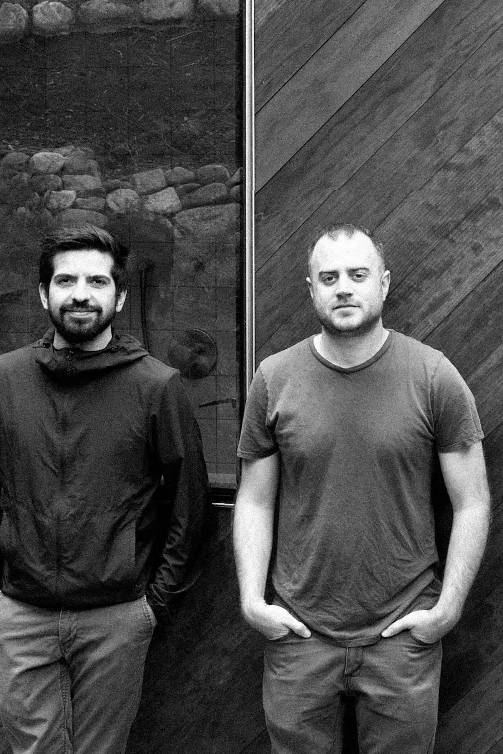 A black and white photograph of two people standing against dark contrasting stone & wooden wooden background. Both are wearing casual attire, one in a long-sleeved shirt , the other in a t-shirt.