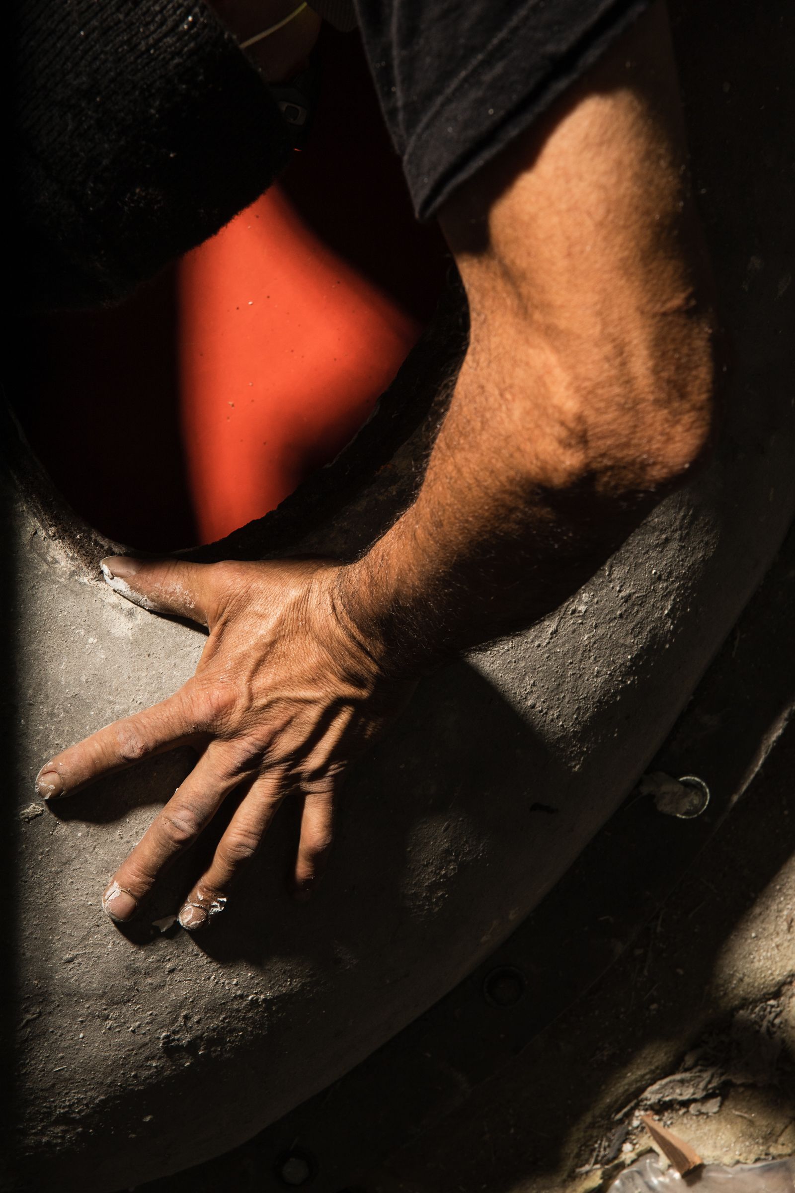 A close-up of a person’s hand pressing against a rough concrete surface, with warm light casting dramatic shadows. The arm extends into a circular mold with a vivid red interior, highlighting the tactile and hands-on nature of concrete craftsmanship.