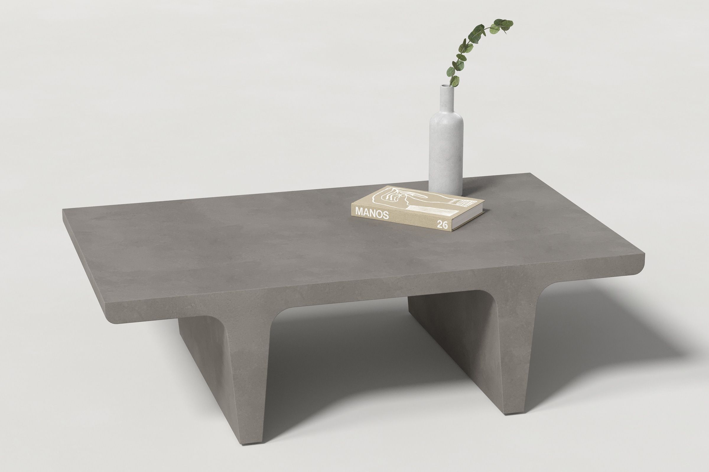 A rectangular dark grey concrete coffee table with sculptural double tee legs, styled with a neutral-toned book and a slender vase holding a eucalyptus, set against a clean white background.