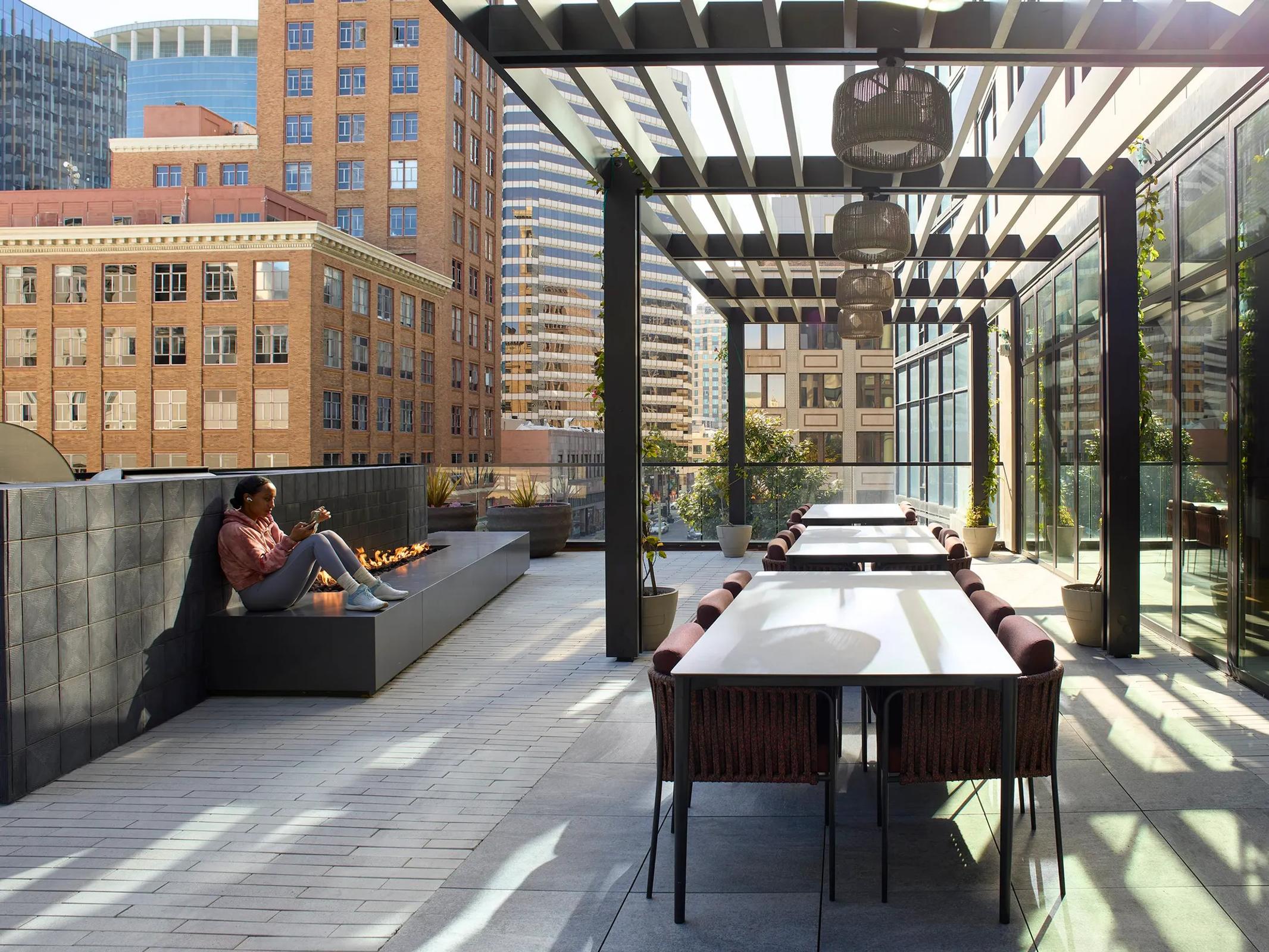 A rooftop dining area with long tables and brown chairs. A person sits on a long fireplace. Hanging woven lamps hang down, with city views of brick buildings in the background.