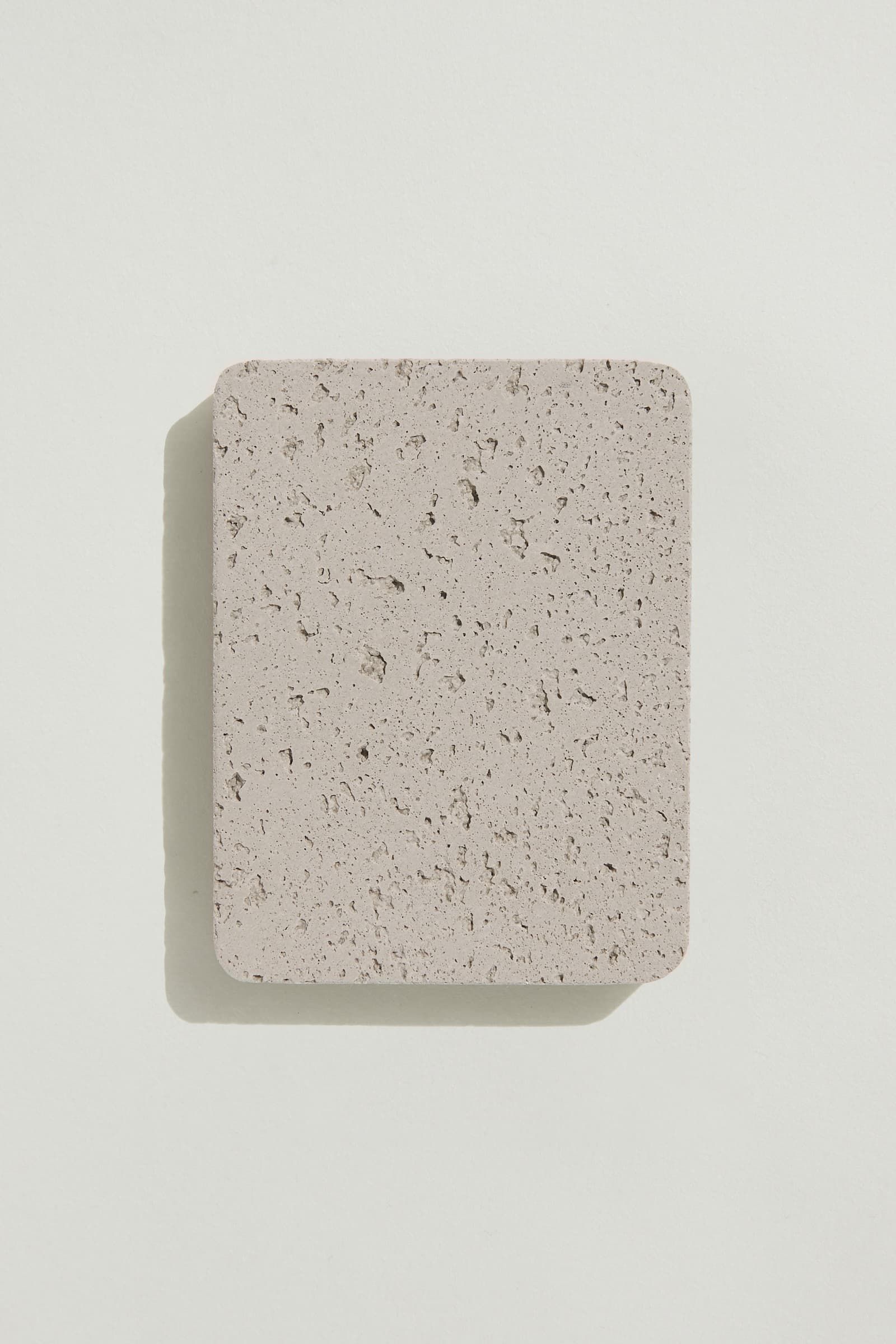 A rectangular sample of rough light grey concrete with a natural, pitted surface texture. Photographed on a white background with a cast shadow.
