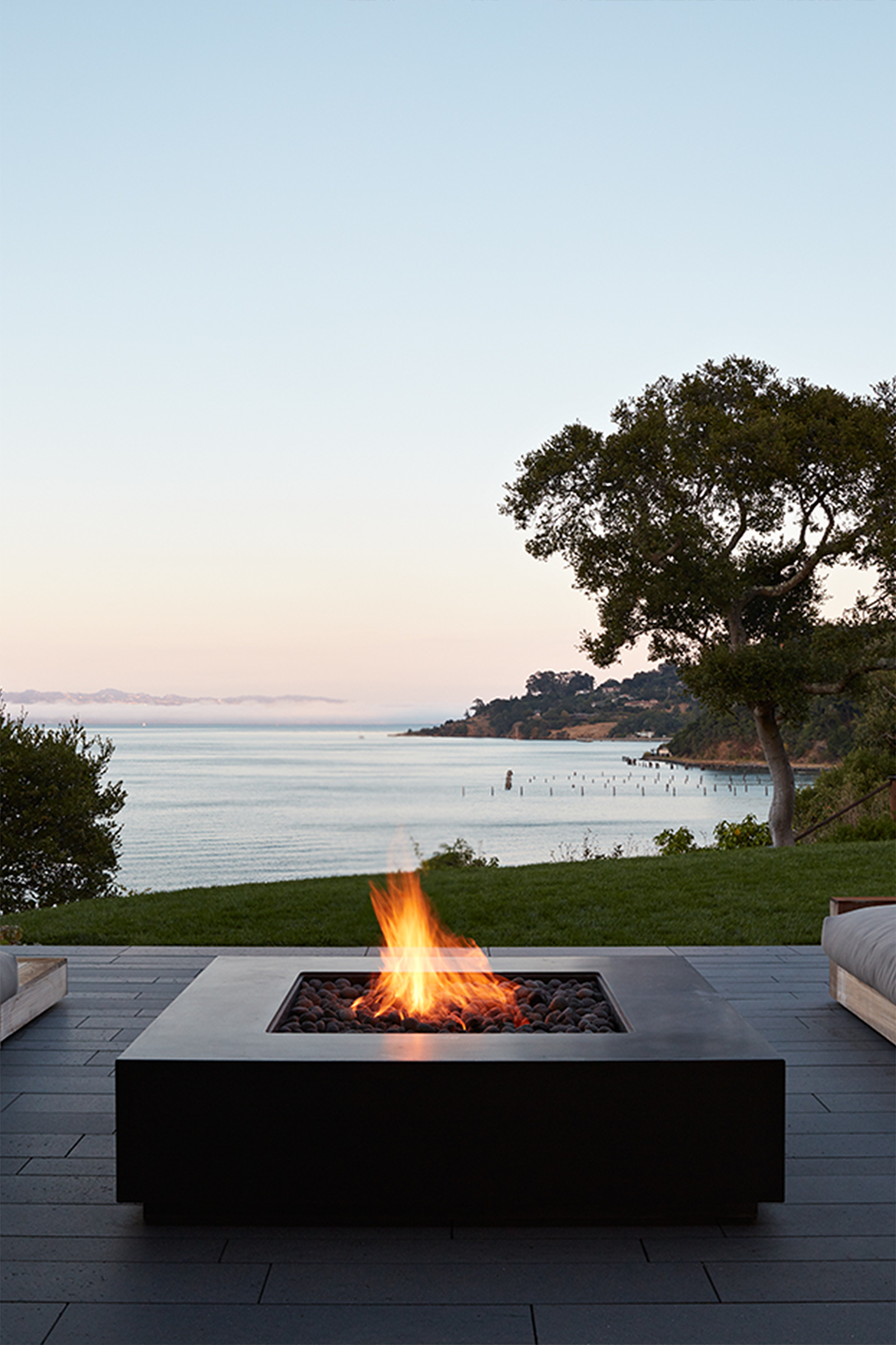 Contemporary waterfront patio with two outdoor sofas facing a dark grey, square concrete fire pit, overlooking a bay at sunset. Framed by trees and soft landscape lighting with a view of the ocean and hills.
