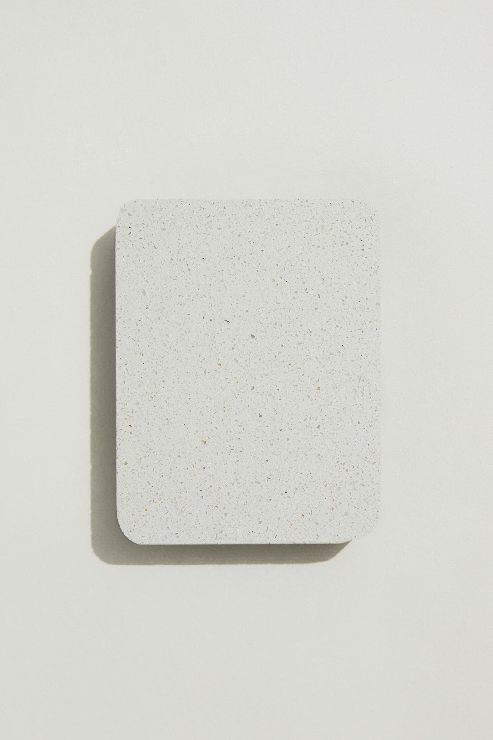 A rectangular sample of smooth white concrete with grey and black terrazzo speckling, and rounded corners. Photographed on a white background with a cast shadow