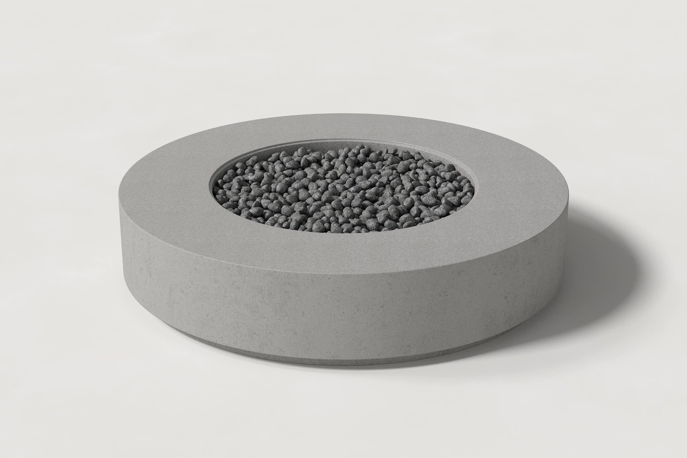Low cylindrical grey concrete fire pit with thick walls and a central circular well. Mid-range angle shows the well filled with volcanic rocks against a white background.