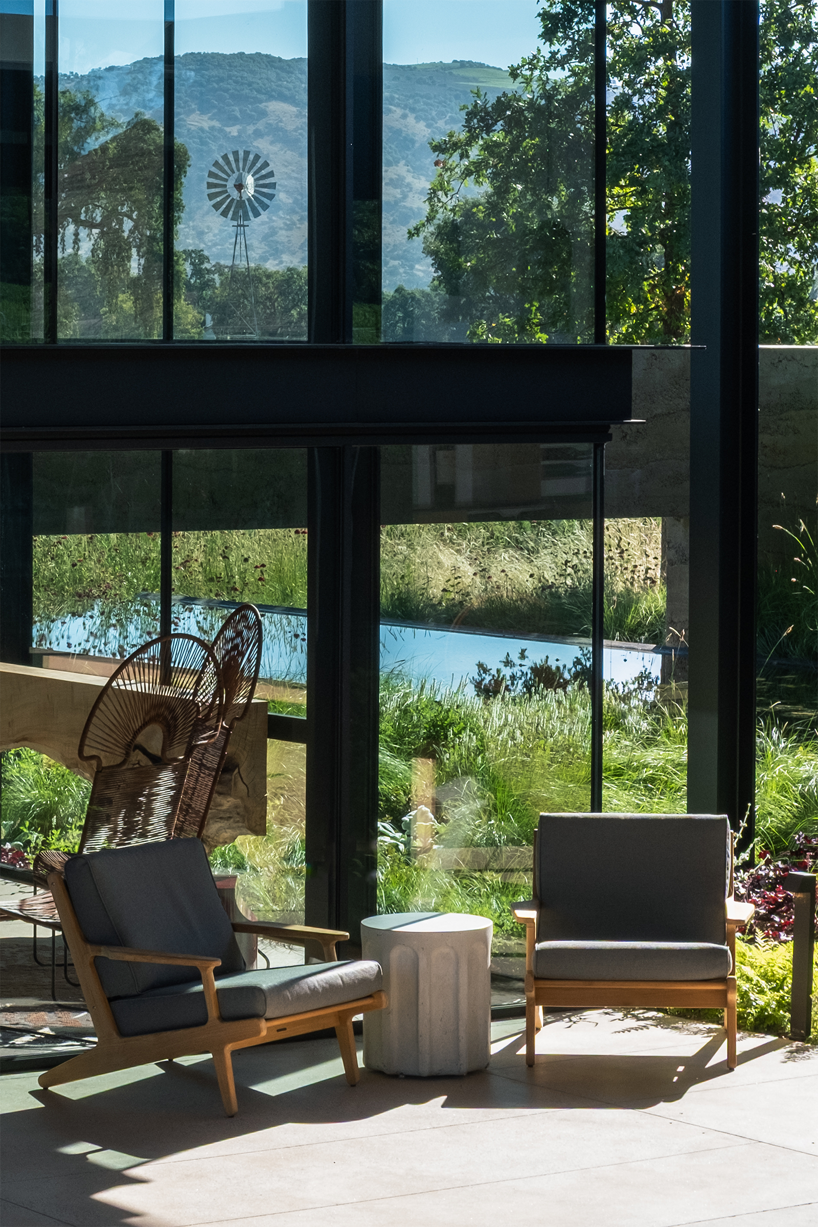 Contemporary outdoor lounge with two wood-framed chairs and a concrete Forum side table overlooking landscaped grounds and distant mountains through floor-to-ceiling windows.