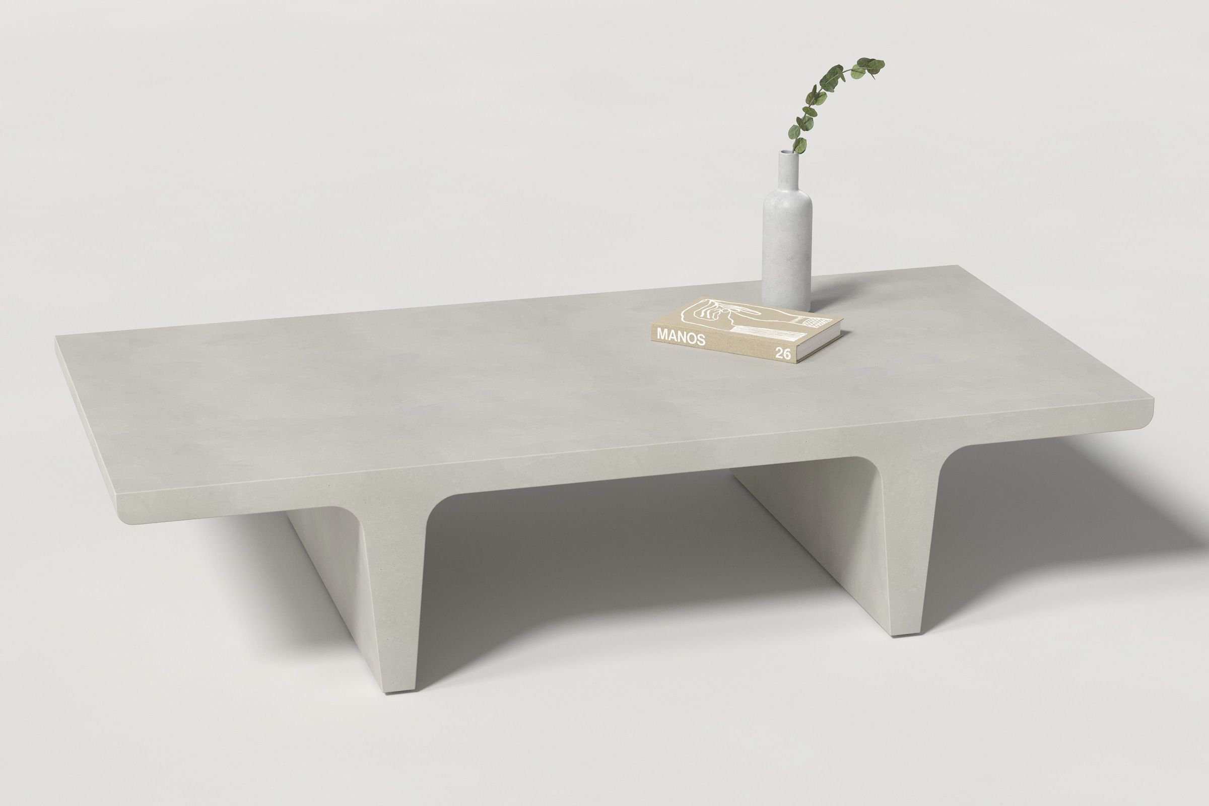 A rectangular light grey concrete coffee table with sculptural double tee legs, styled with a neutral-toned book and a slender vase holding a eucalyptus, set against a clean white background.