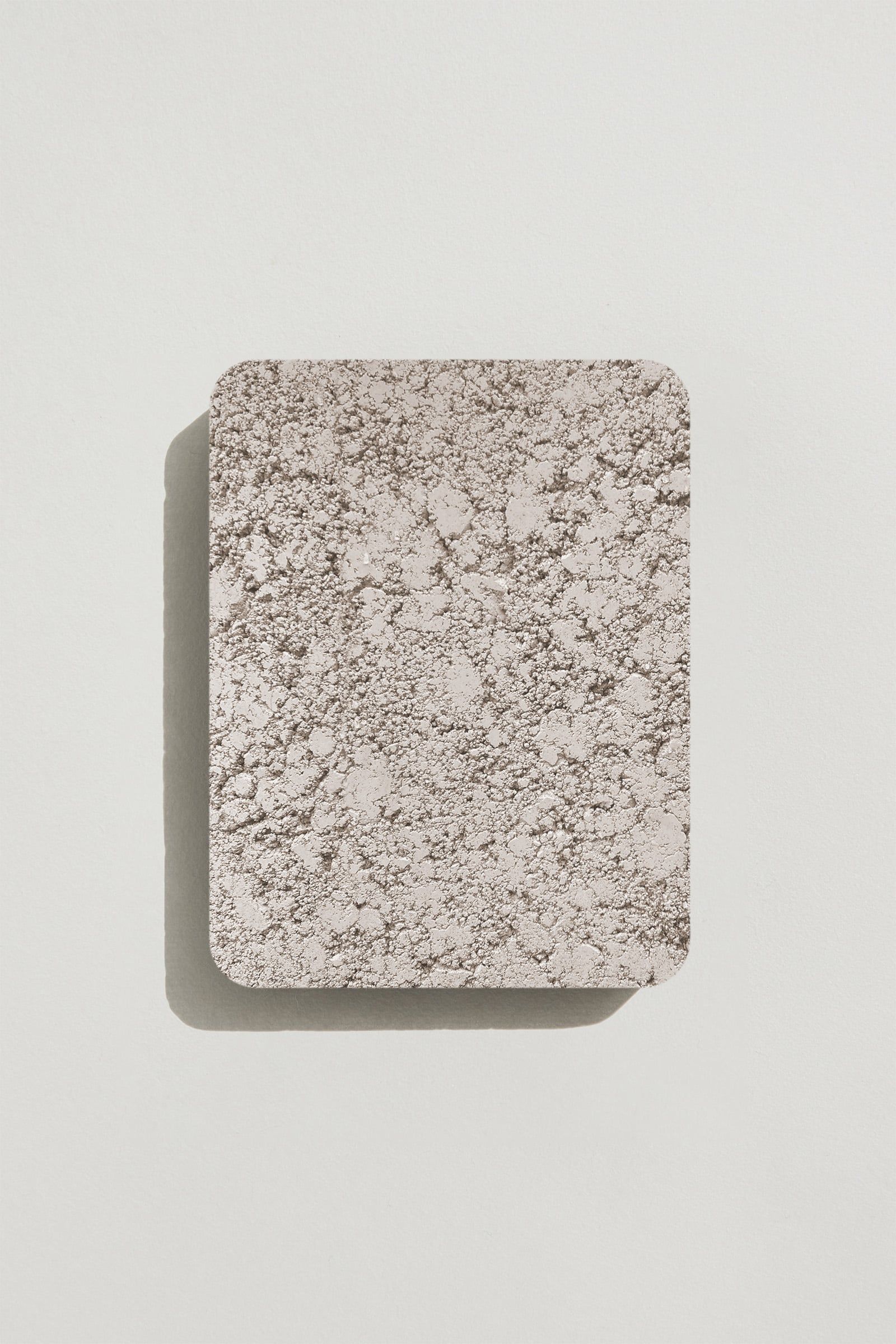 A rectangular sample of rough light grey concrete with a natural, pitted surface texture. Photographed on a white background with a cast shadow.