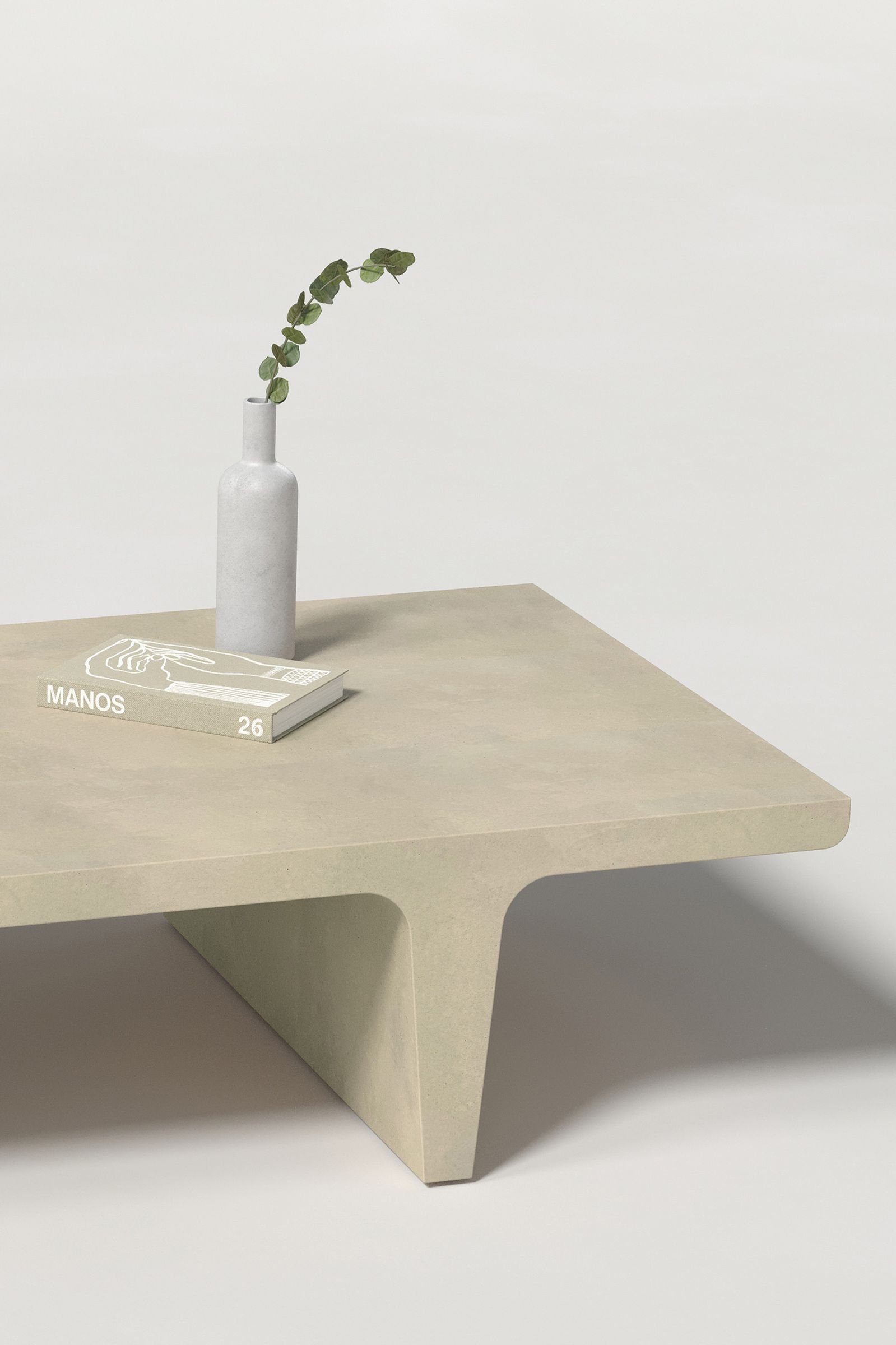 Close-up of a rectangular light beige concrete coffee table with sculptural double tee legs, styled with a neutral-toned book and a slender vase holding a eucalyptus, set against a clean white background.